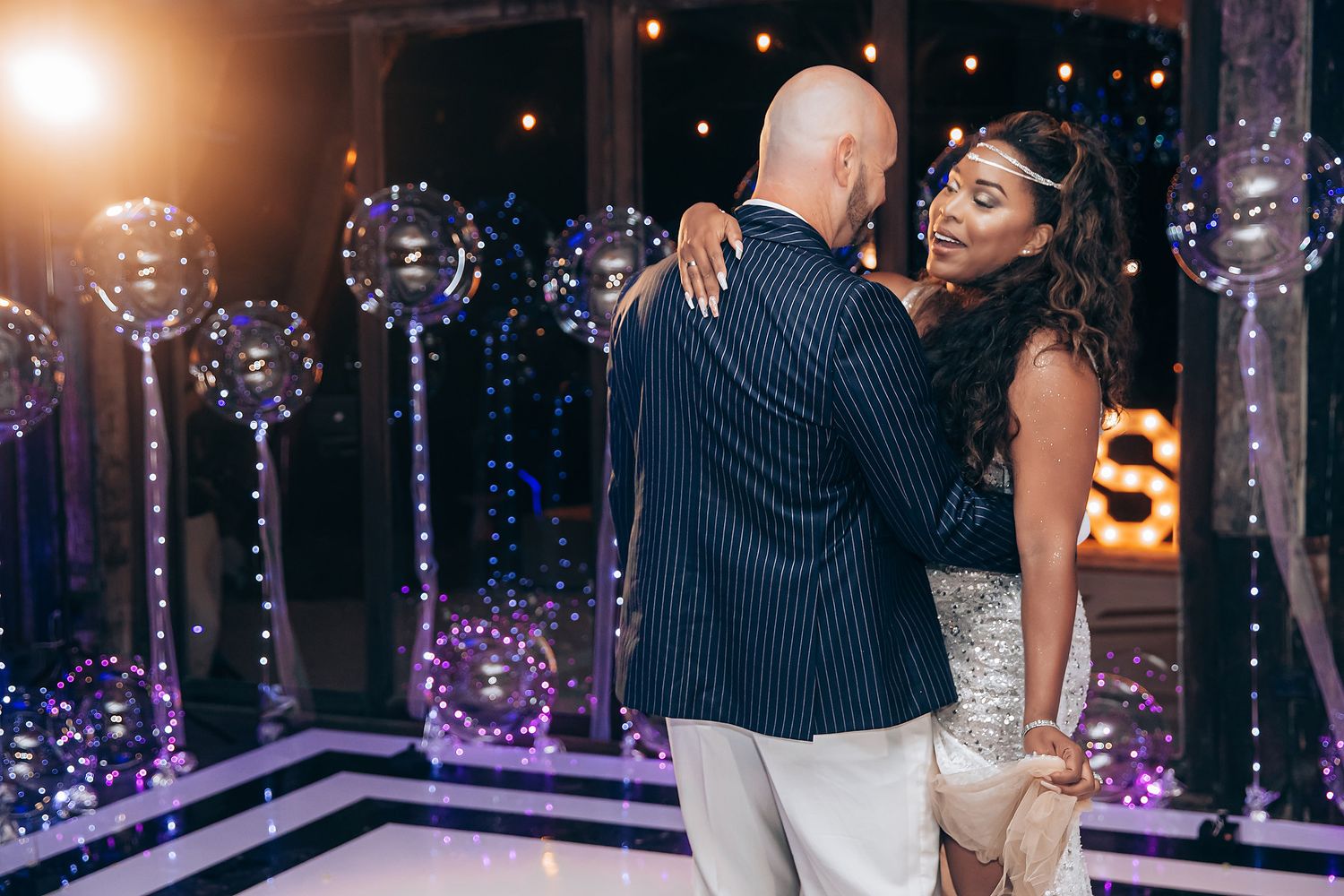 A couple dances intimately on a dance floor decorated with glowing purple lights and illuminated balloons.
