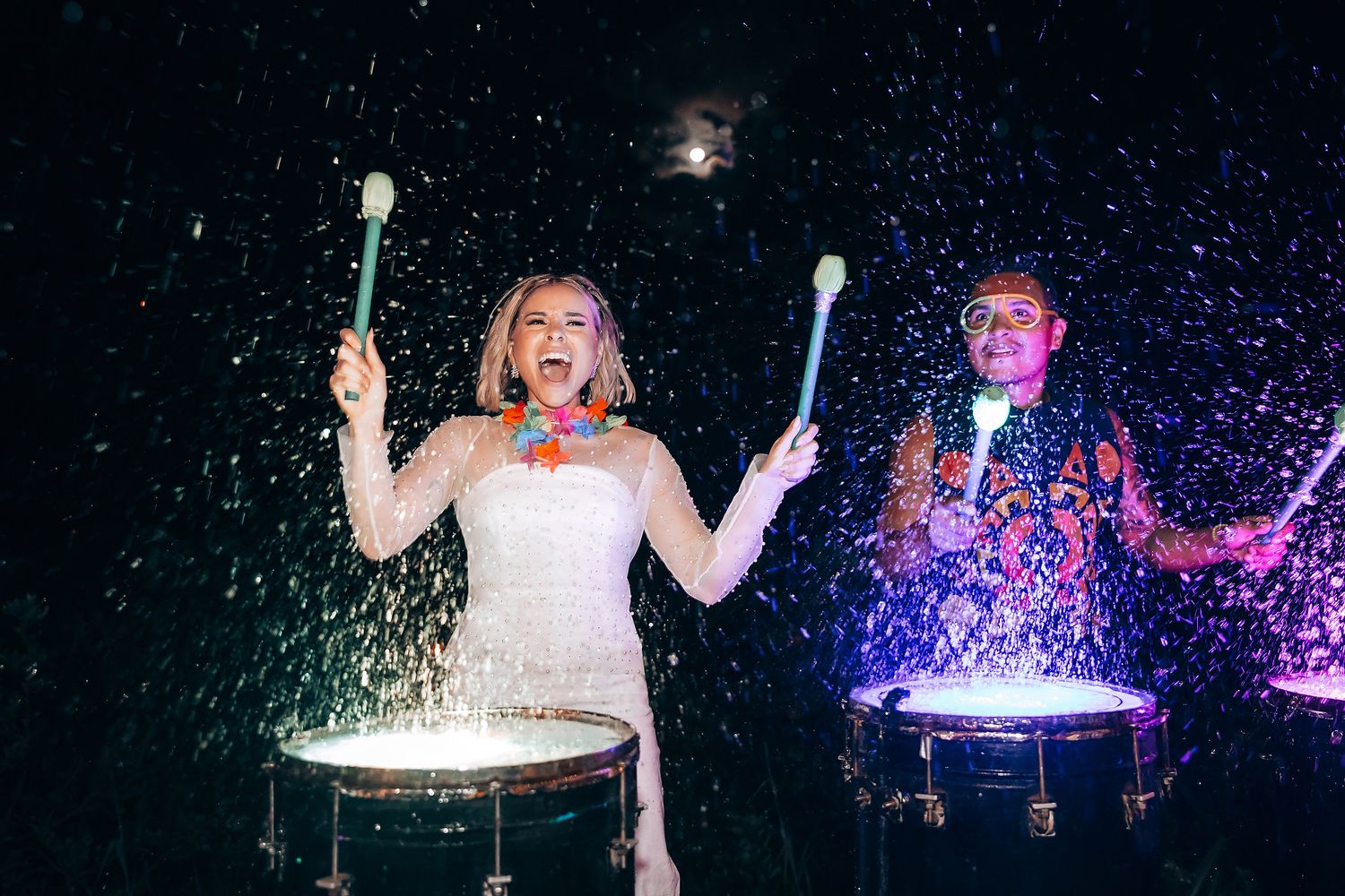 Wedding celebration with couple playing drums amid colorful light show and water splashes.