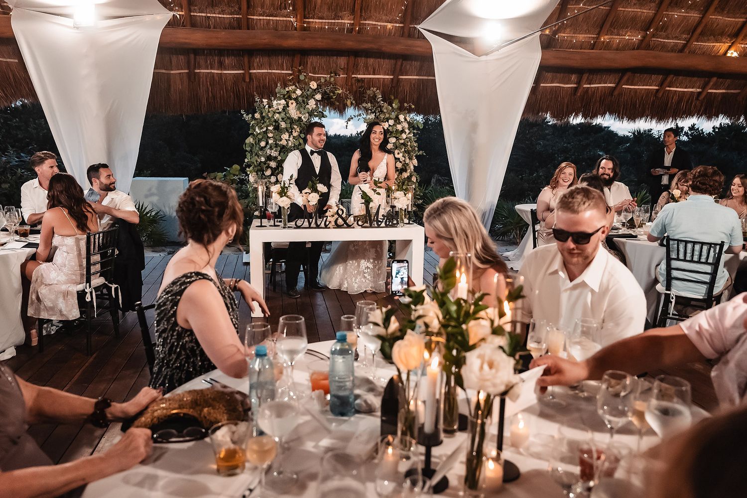 Wedding guests enjoying a reception dinner under white drapes in a rustic barn venue with warm lighting.