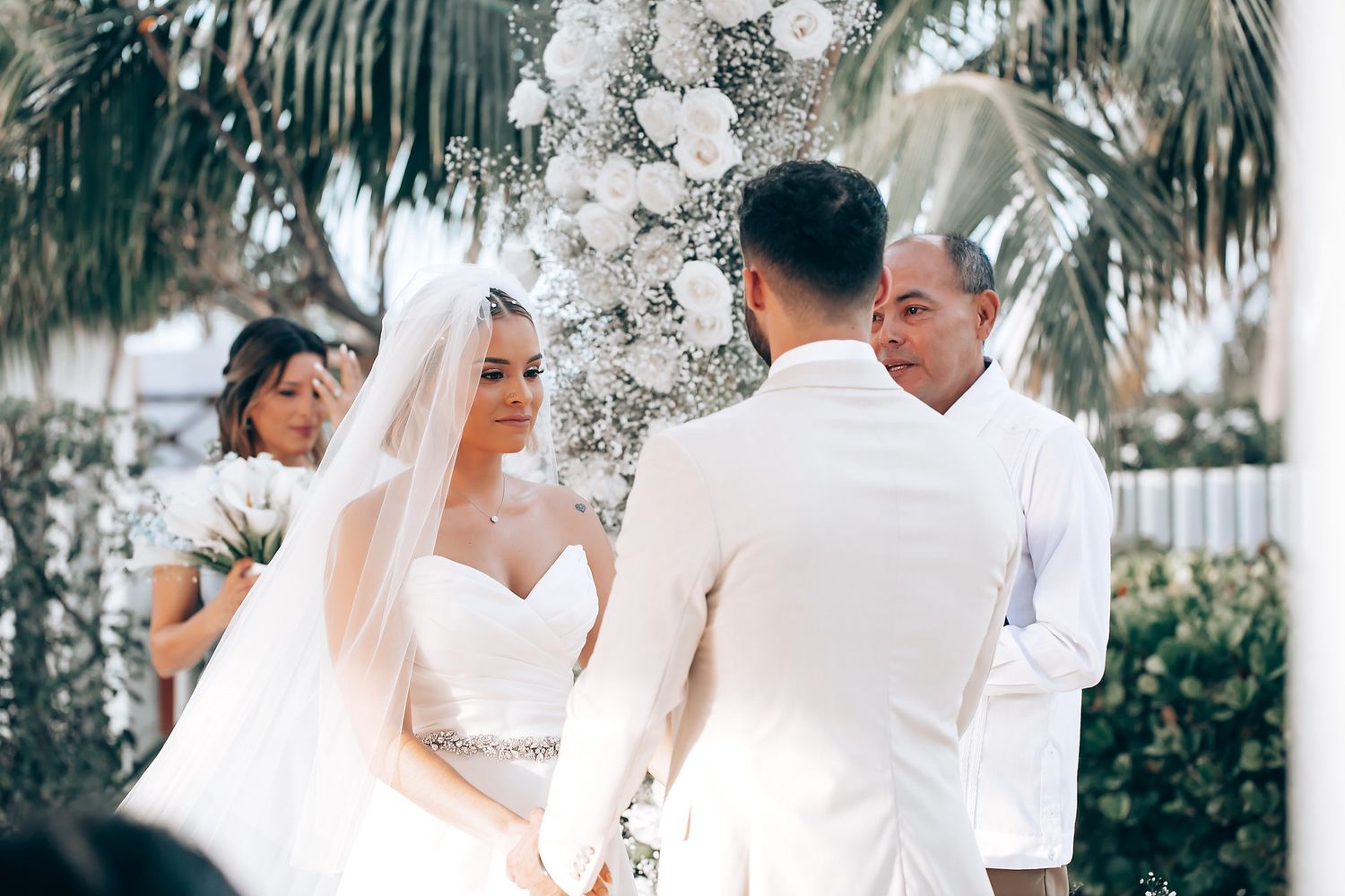 Elegant outdoor wedding ceremony setup with white floral arrangements and palm trees in the background.