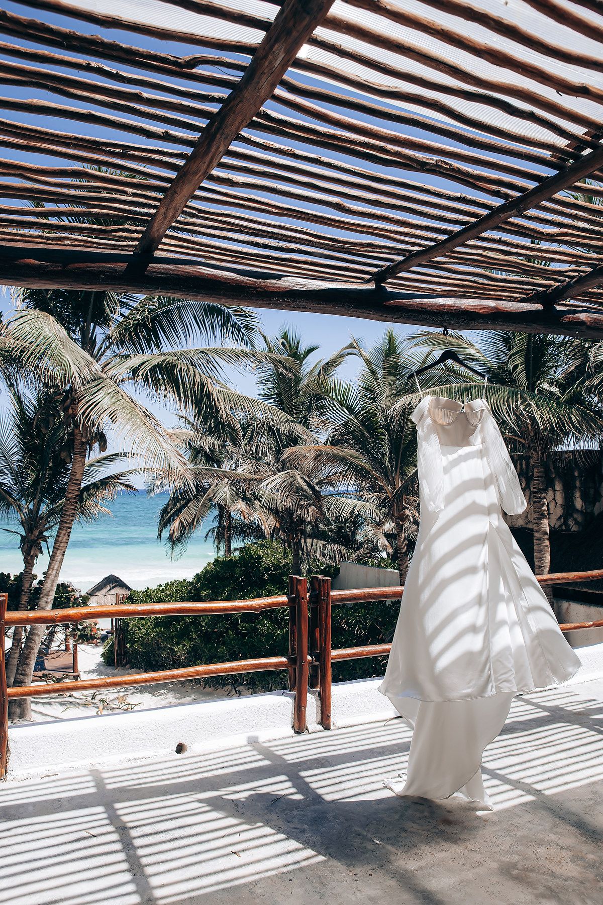 White wedding dress flowing in the breeze under a wooden pergola with palm trees and ocean views.