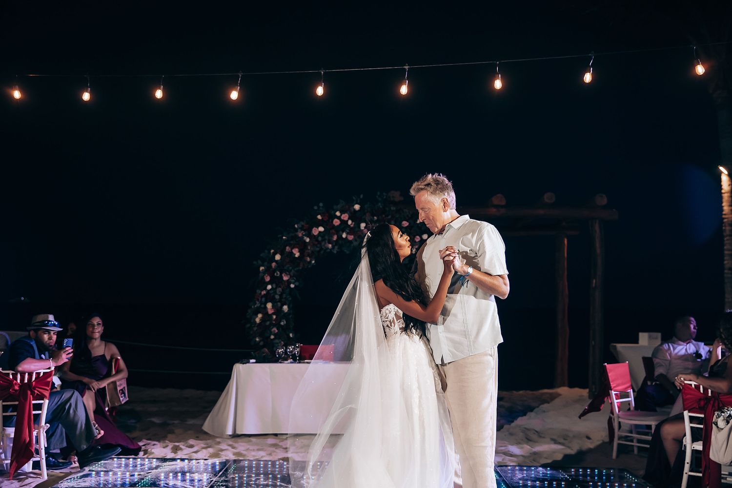 Evening wedding reception under string lights with dancing silhouettes by the beach.