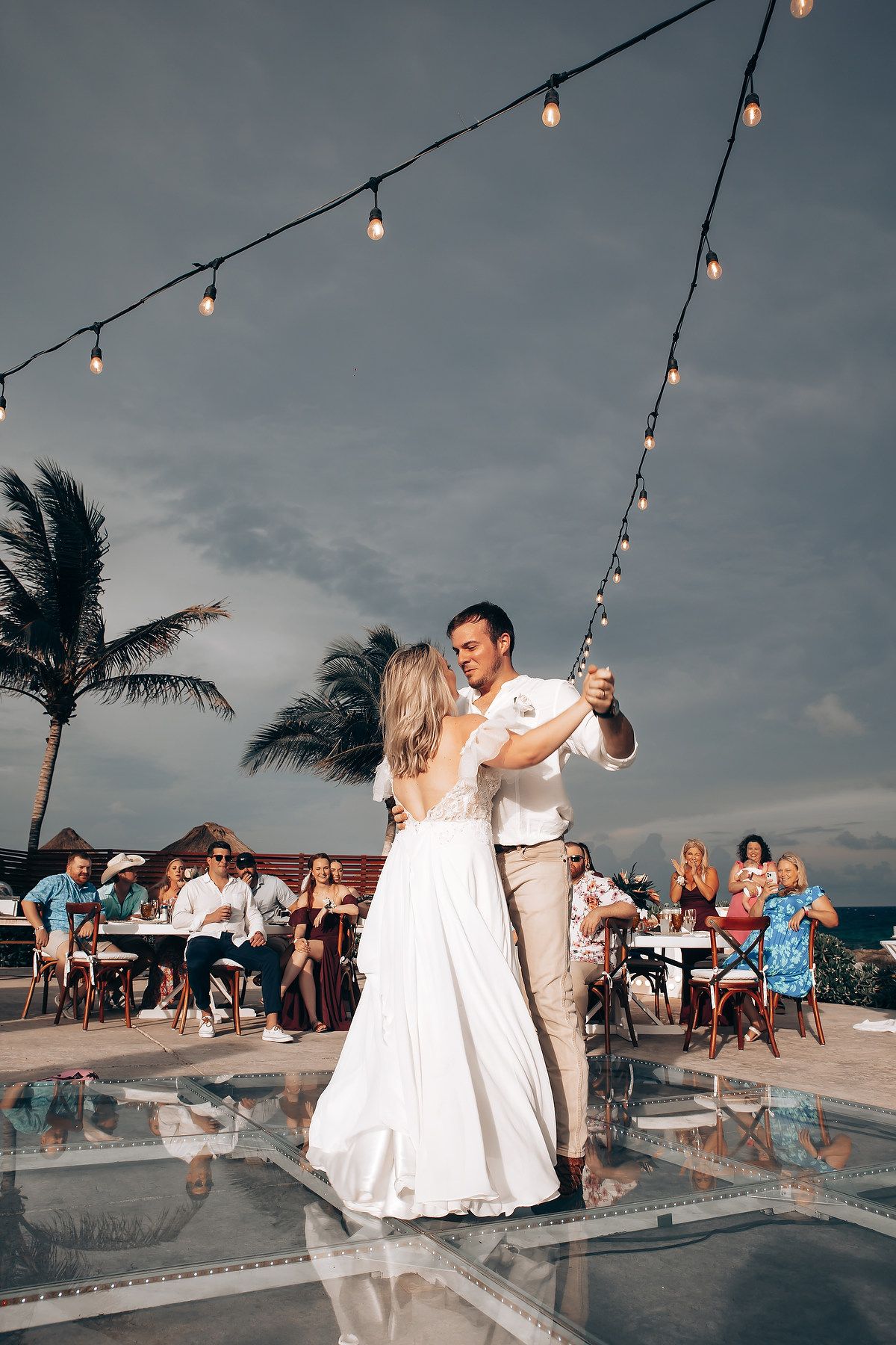 Evening reception dance under string lights with palm trees and ocean backdrop.