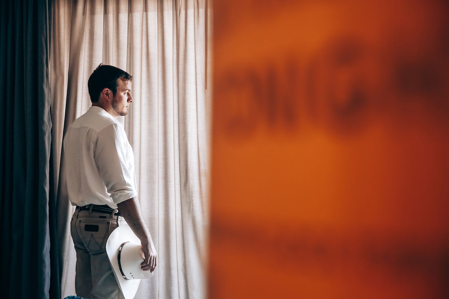 Silhouette of person in white shirt standing by window with orange wall and curtains.