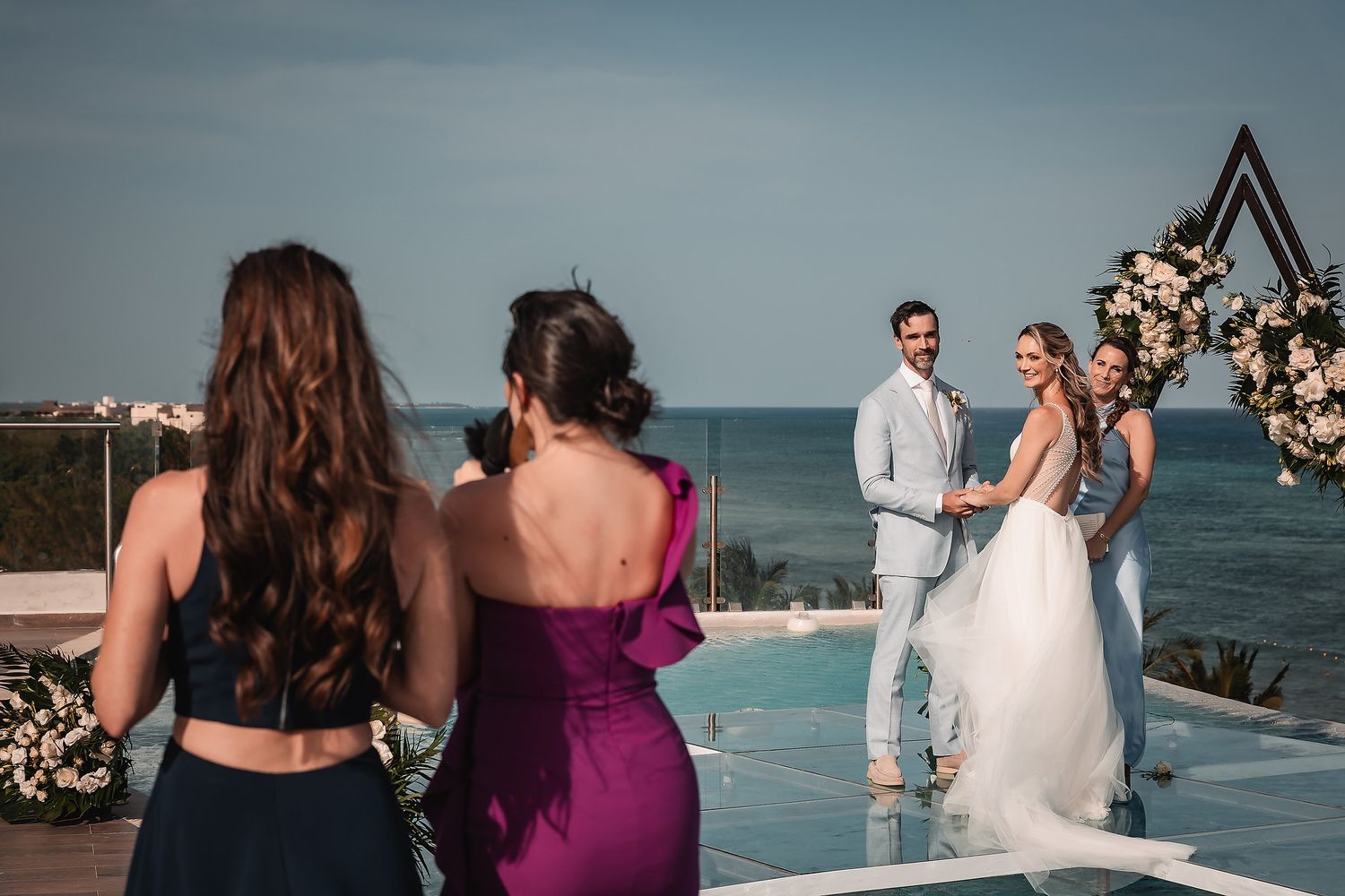 Wedding ceremony on an oceanfront deck with guests watching a couple exchange vows under a floral arch.