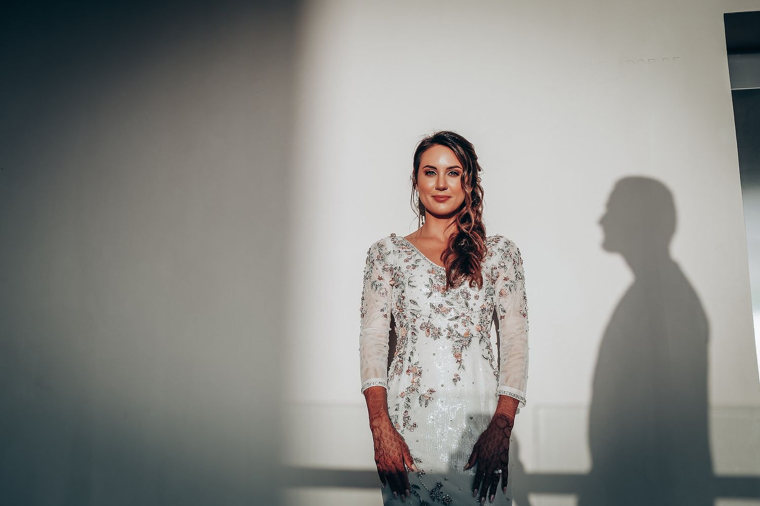 A dramatic portrait against a white wall showing an elegant lace wedding dress with a shadow cast to the side.