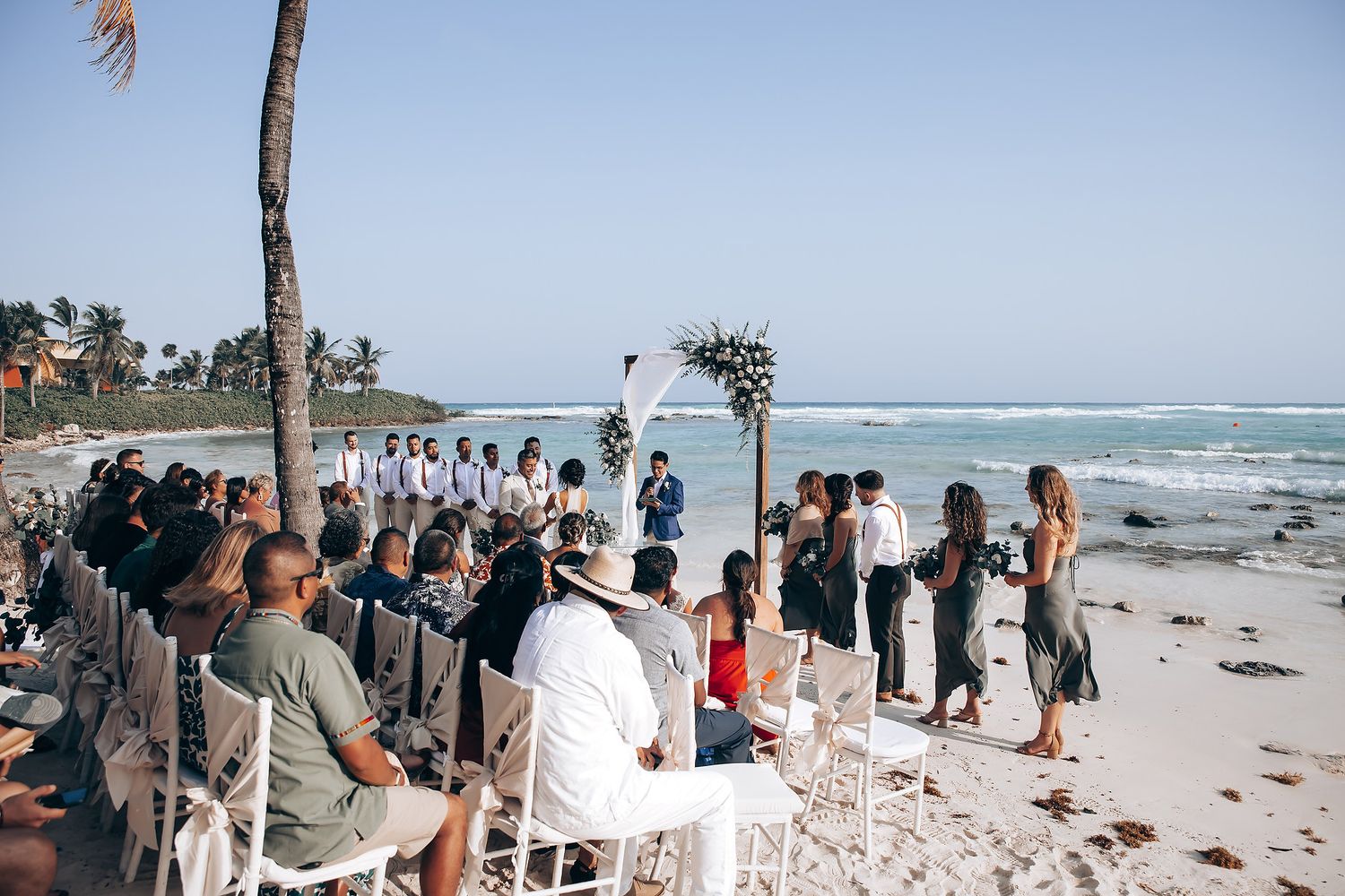 Guests seated in white chairs watch a beachside wedding ceremony with palm trees and ocean views.