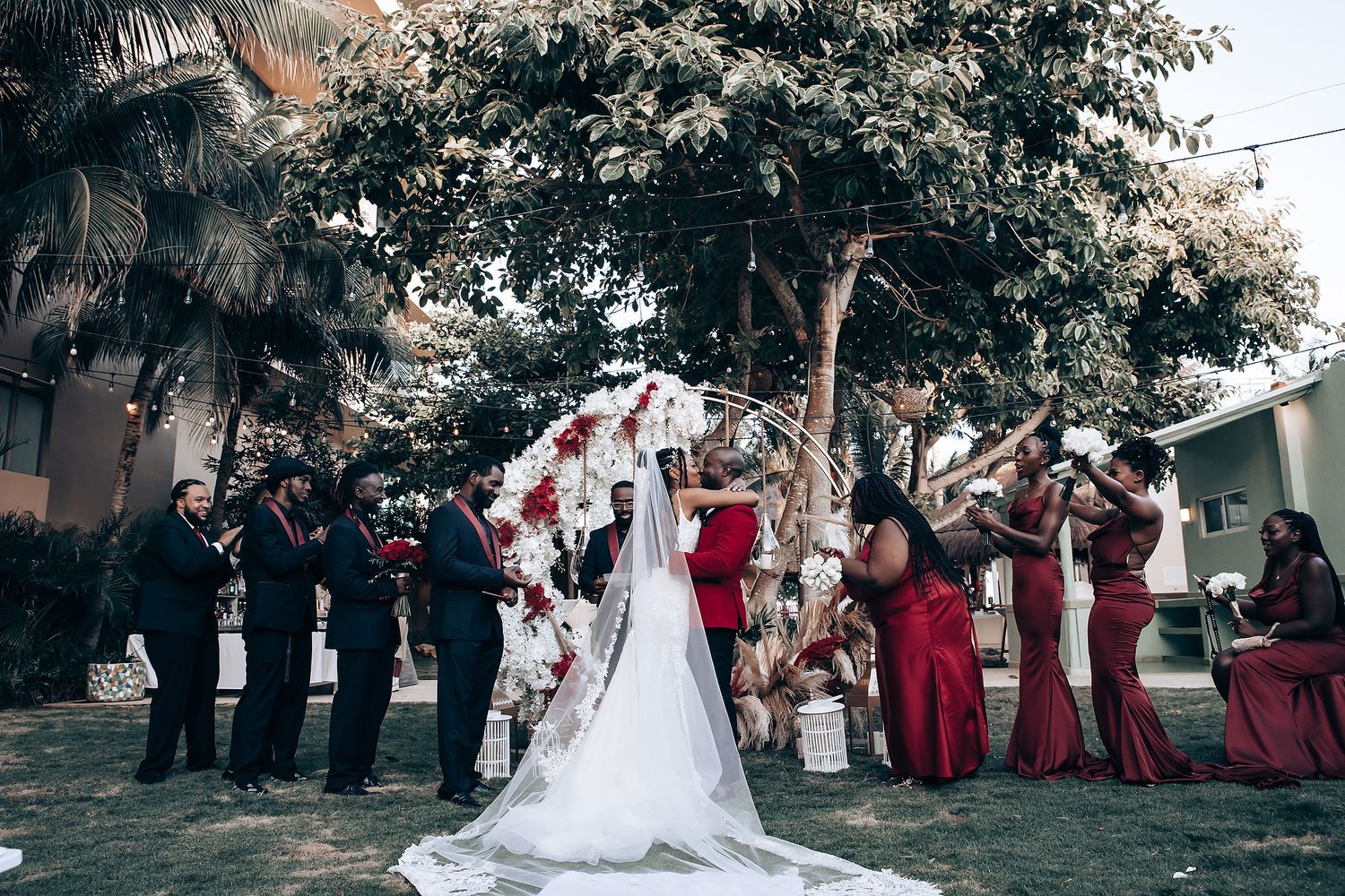 Wedding party poses under trees with bridesmaids in burgundy dresses and bride in white gown.