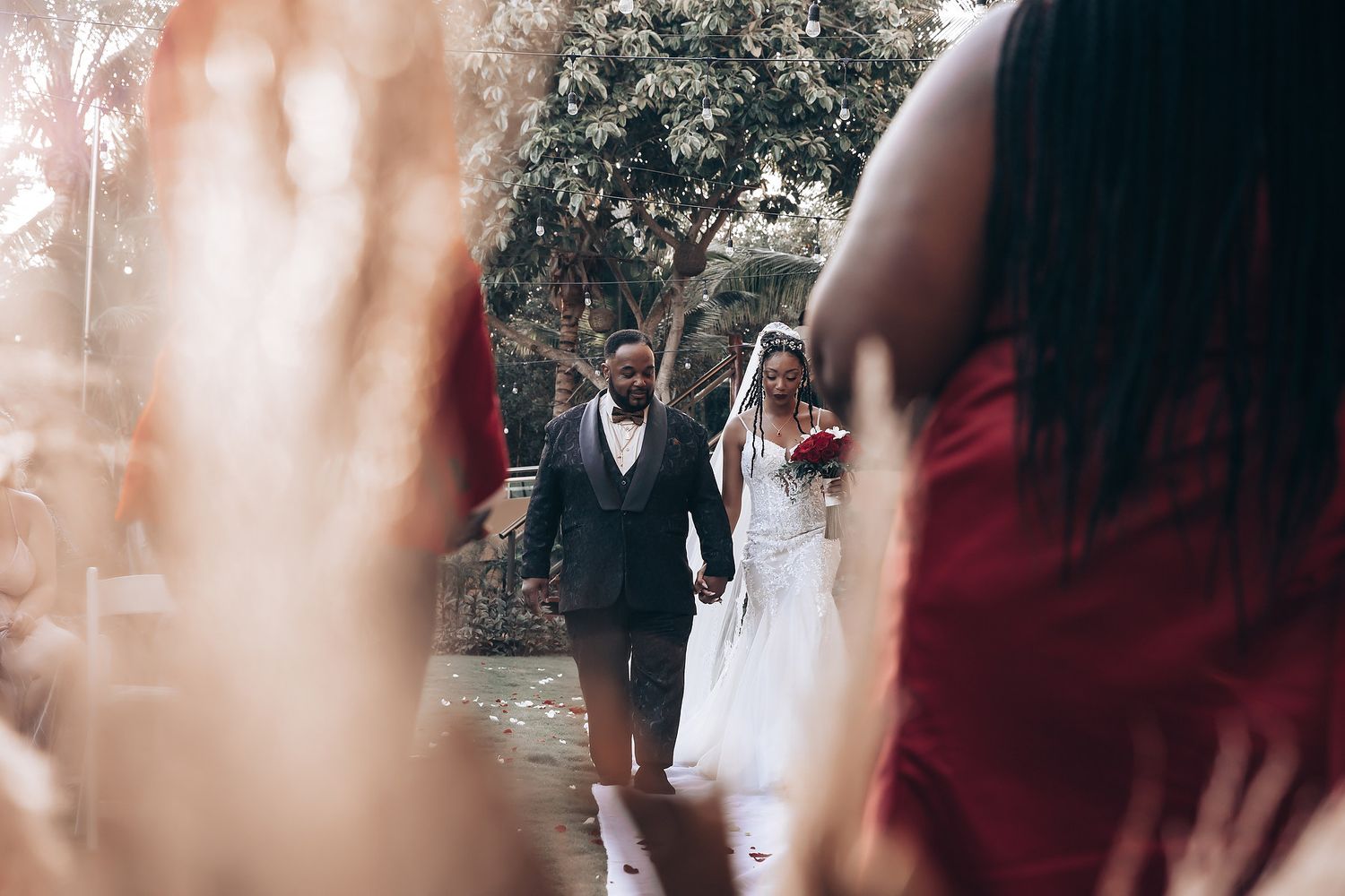 Dreamy outdoor wedding scene with guests and red bridesmaid dresses visible through a light haze.