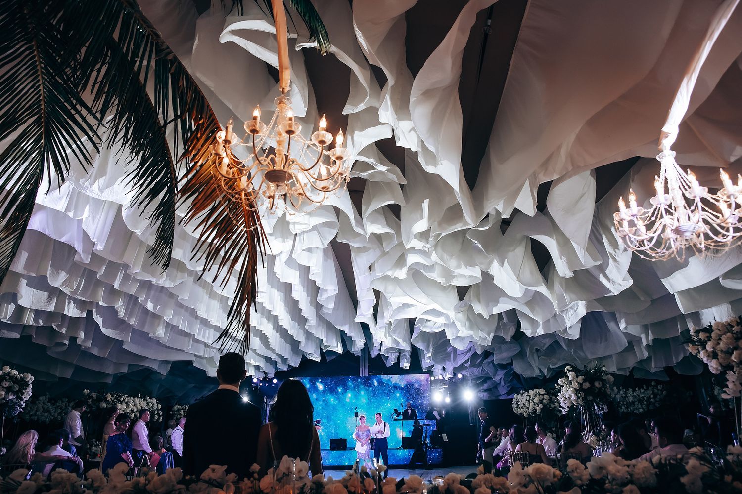 Wedding reception venue with dramatic white fabric ceiling draping and chandeliers.