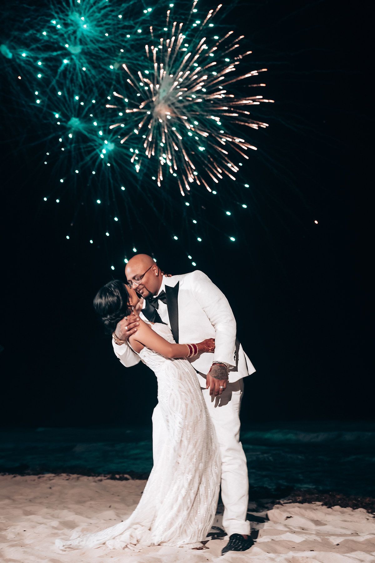 Romantic beach wedding photo with fireworks lighting up night sky in turquoise and white.