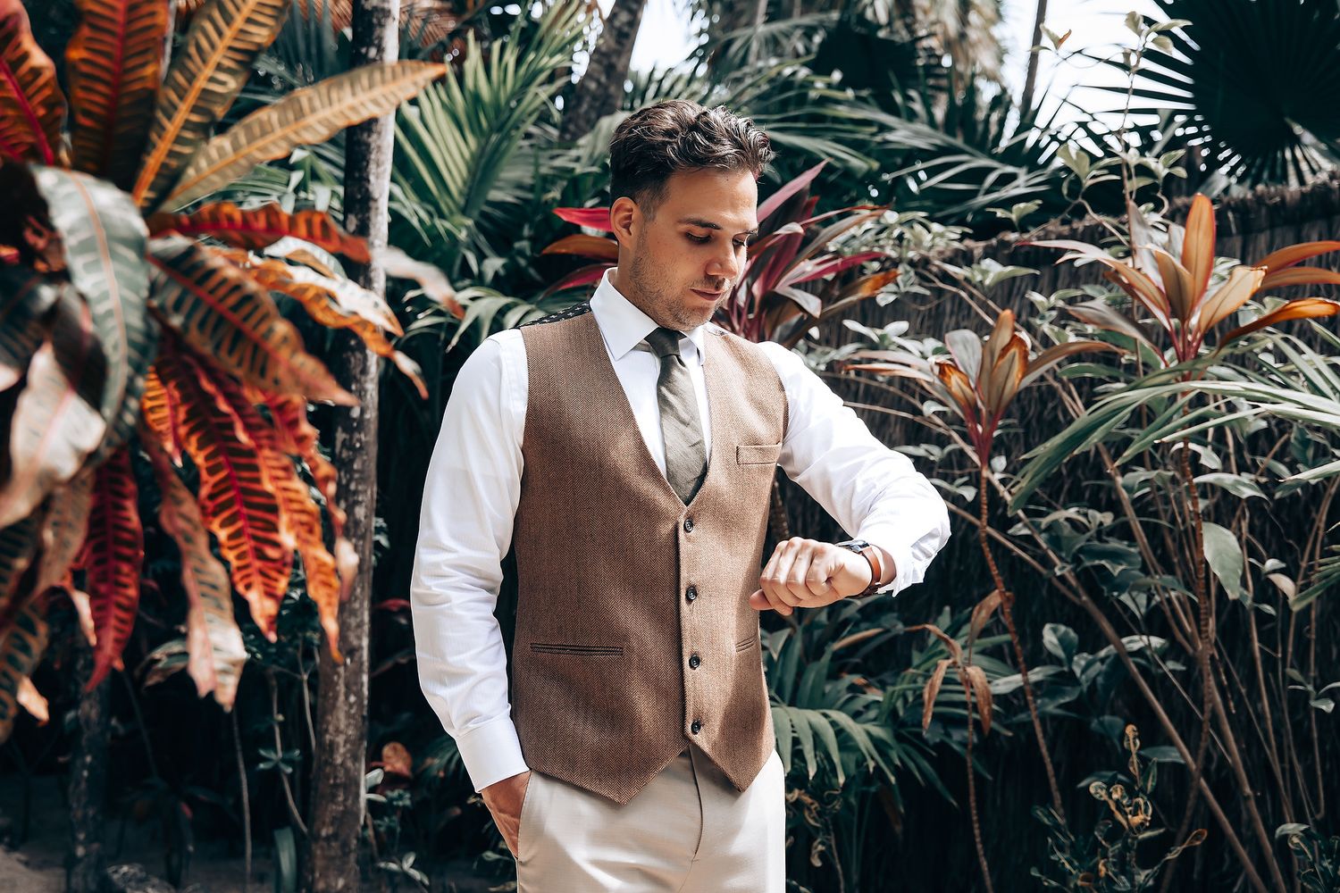 Person in brown vest and white shirt checks time while standing among tropical plants.