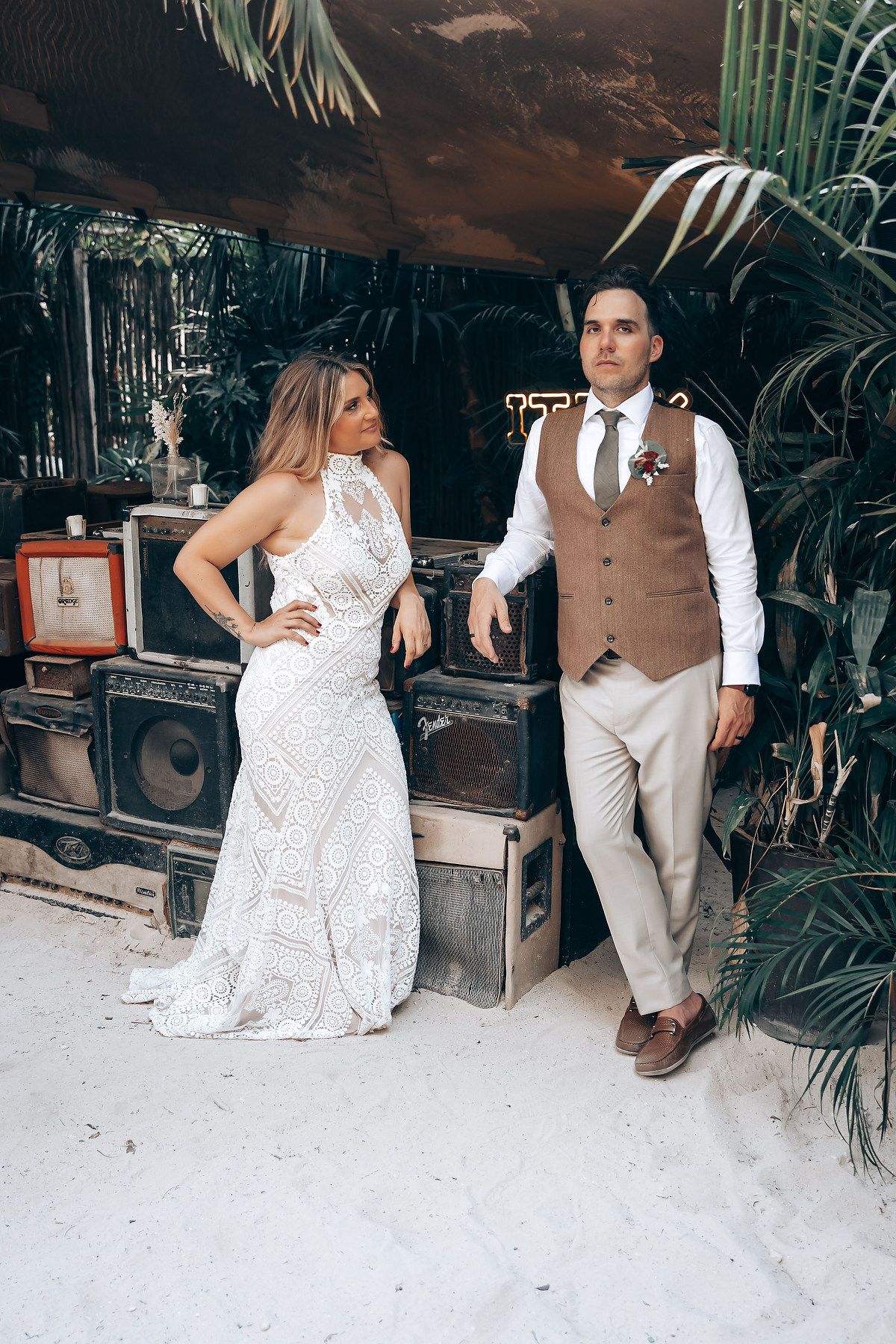 A couple poses in a tropical setting, with her in a white lace dress and him in brown vest and khaki pants.
