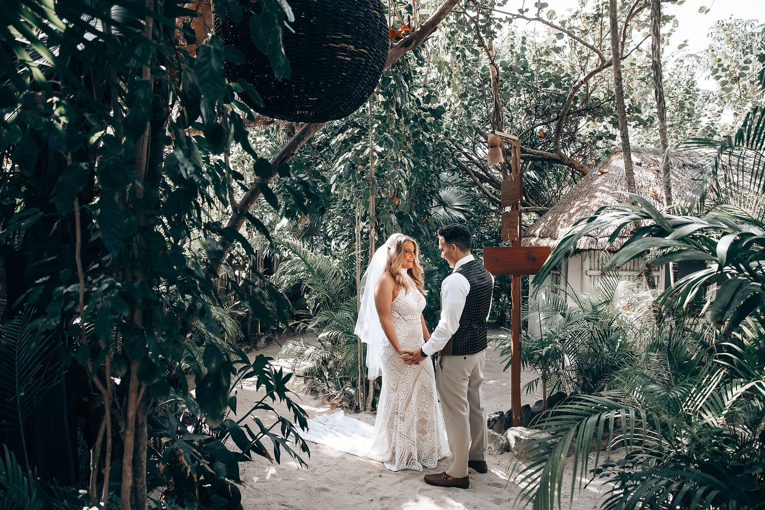A couple in white attire shares an intimate moment in a lush tropical garden setting during their wedding celebration.