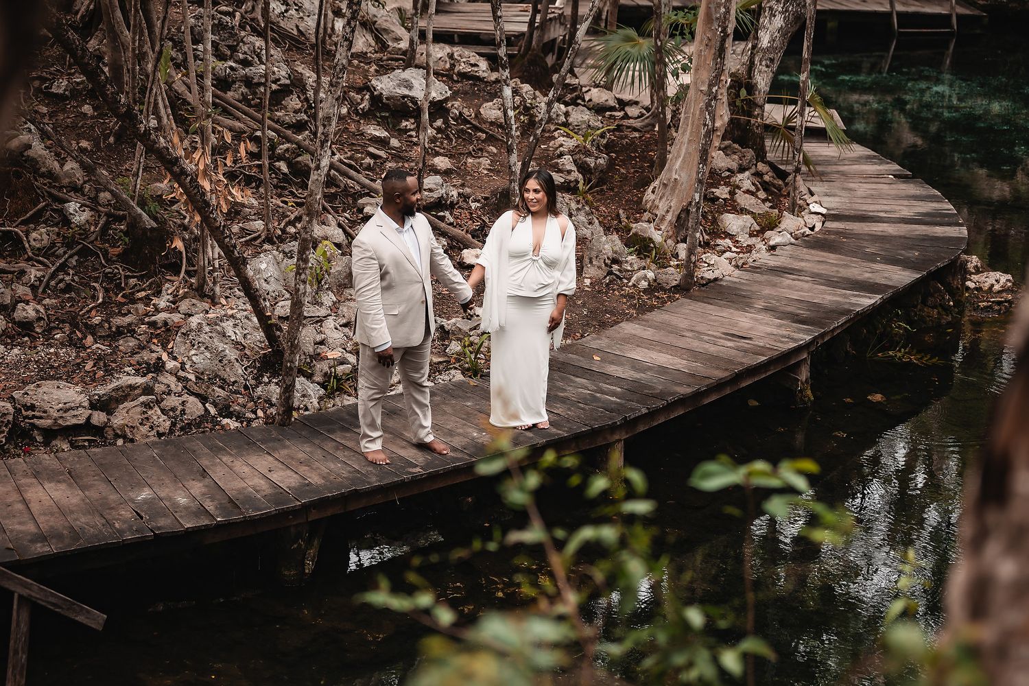 Couple walking along wooden pathway over tranquil cenote waters surrounded by limestone walls.