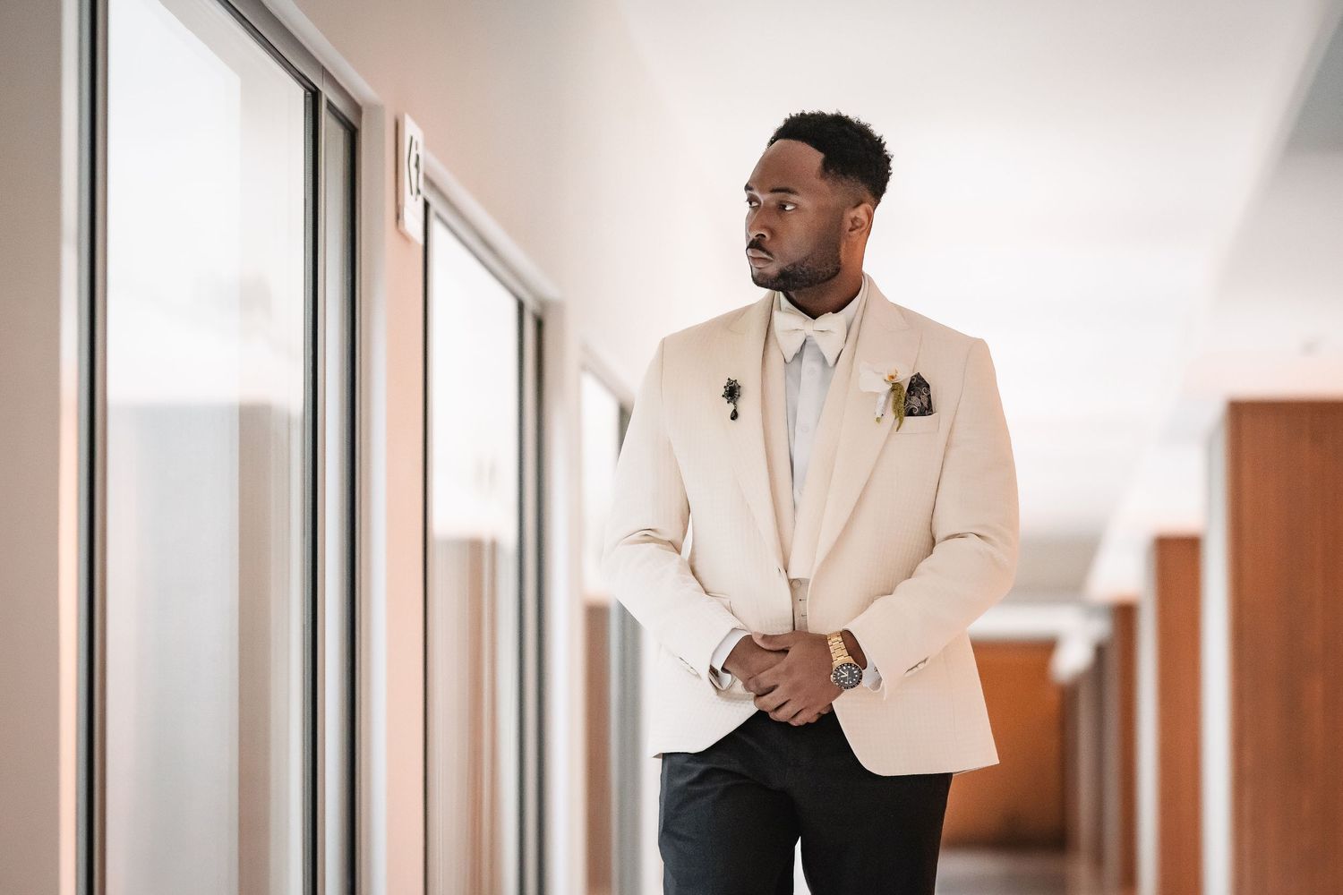A person in a cream-colored suit stands in a modern hallway with glass walls.