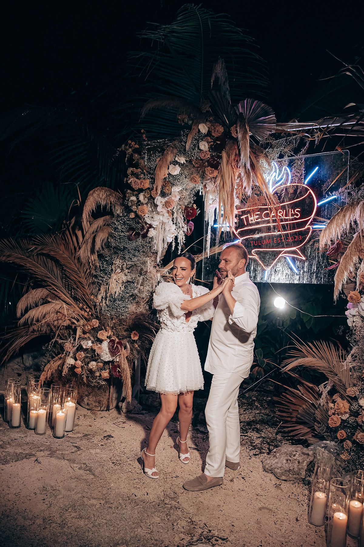 A couple dances at a romantic outdoor venue decorated with pampas grass, candles and a neon sign at night.