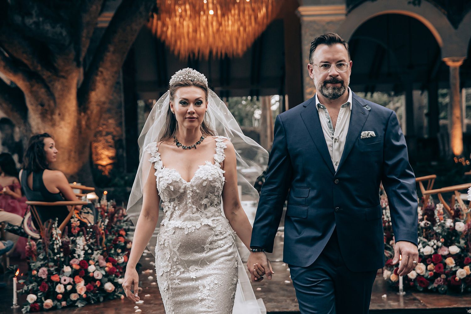 A couple walks together during their wedding ceremony in an elegant indoor venue with warm lighting and floral decor.