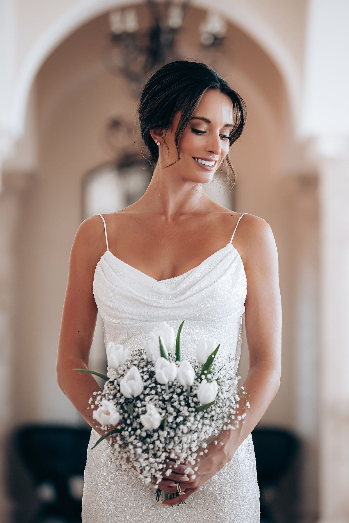 Bride in white spaghetti strap dress holds delicate bouquet of white flowers and baby's breath against light backdrop.