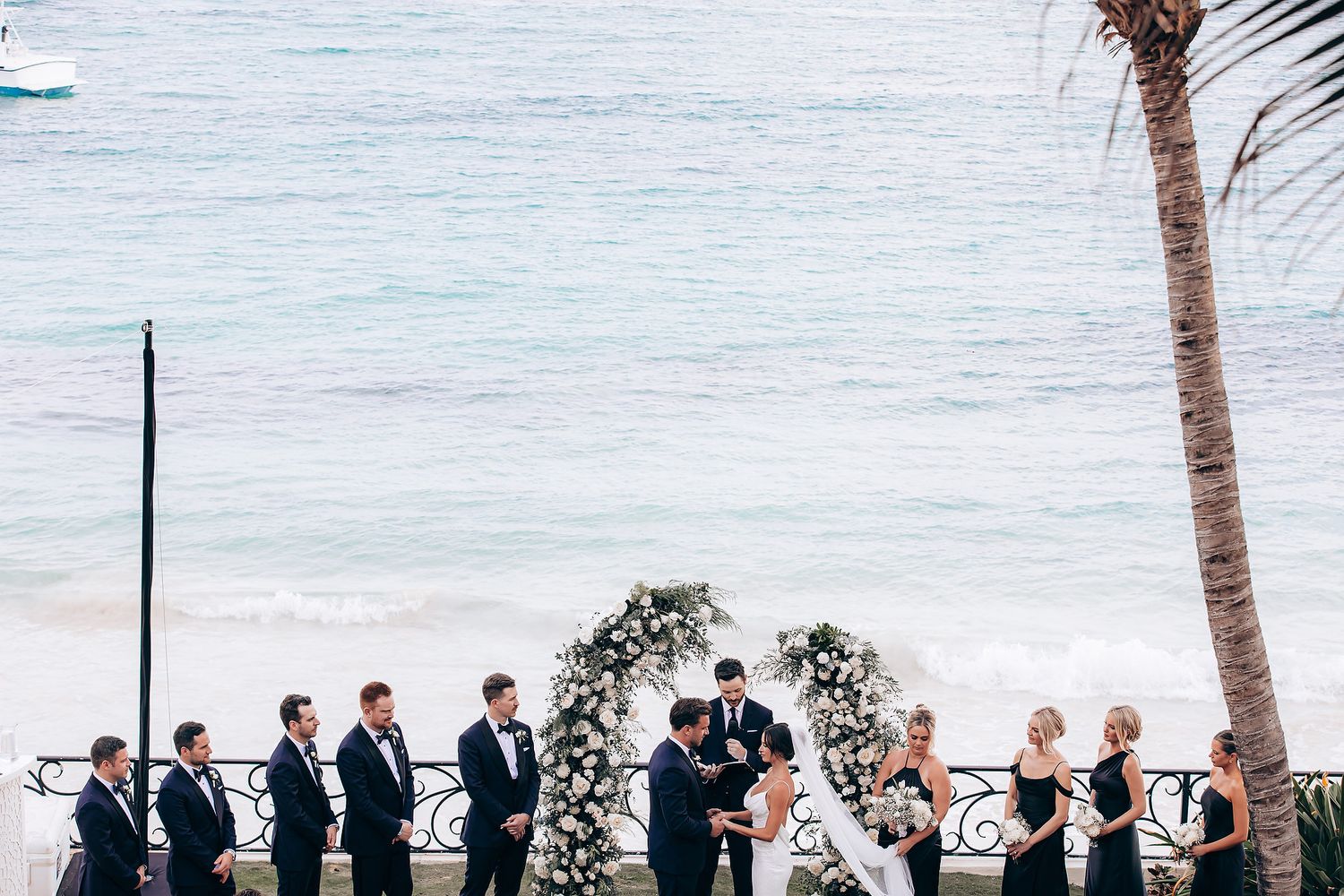 Wedding ceremony setup on a tropical beach with white chairs and floral arch under palm trees at sunset.