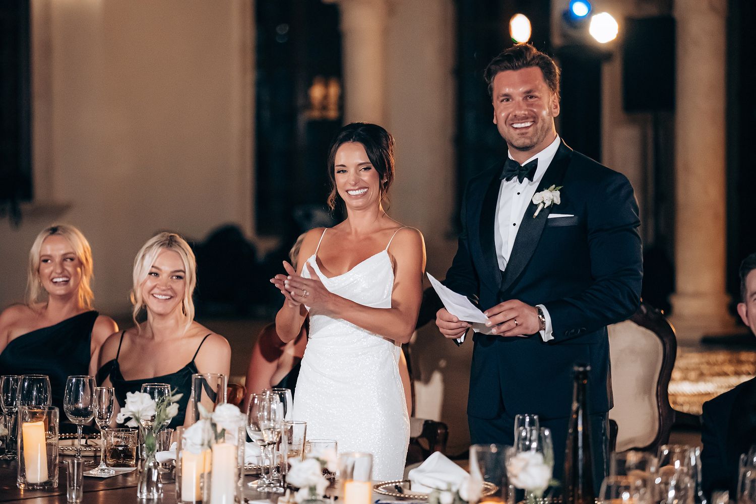 Wedding reception guests seated at elegant banquet tables with glowing candles and floral centerpieces in grand ballroom.