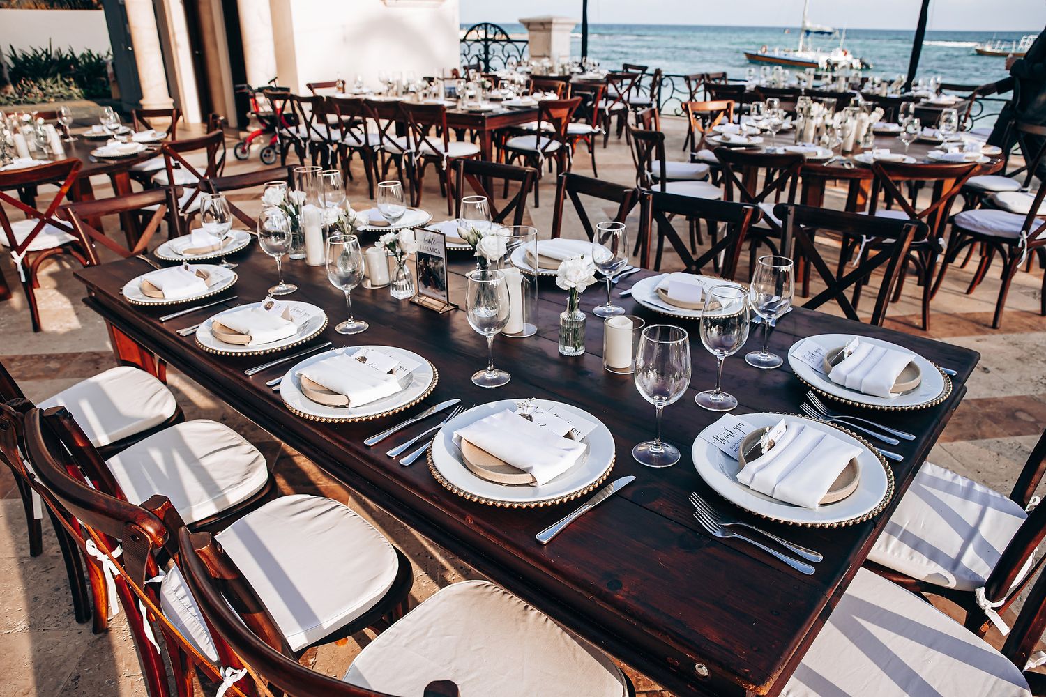 Elegant wedding reception tables set with white plates, crystal glasses and dark wood chairs in a rustic venue.