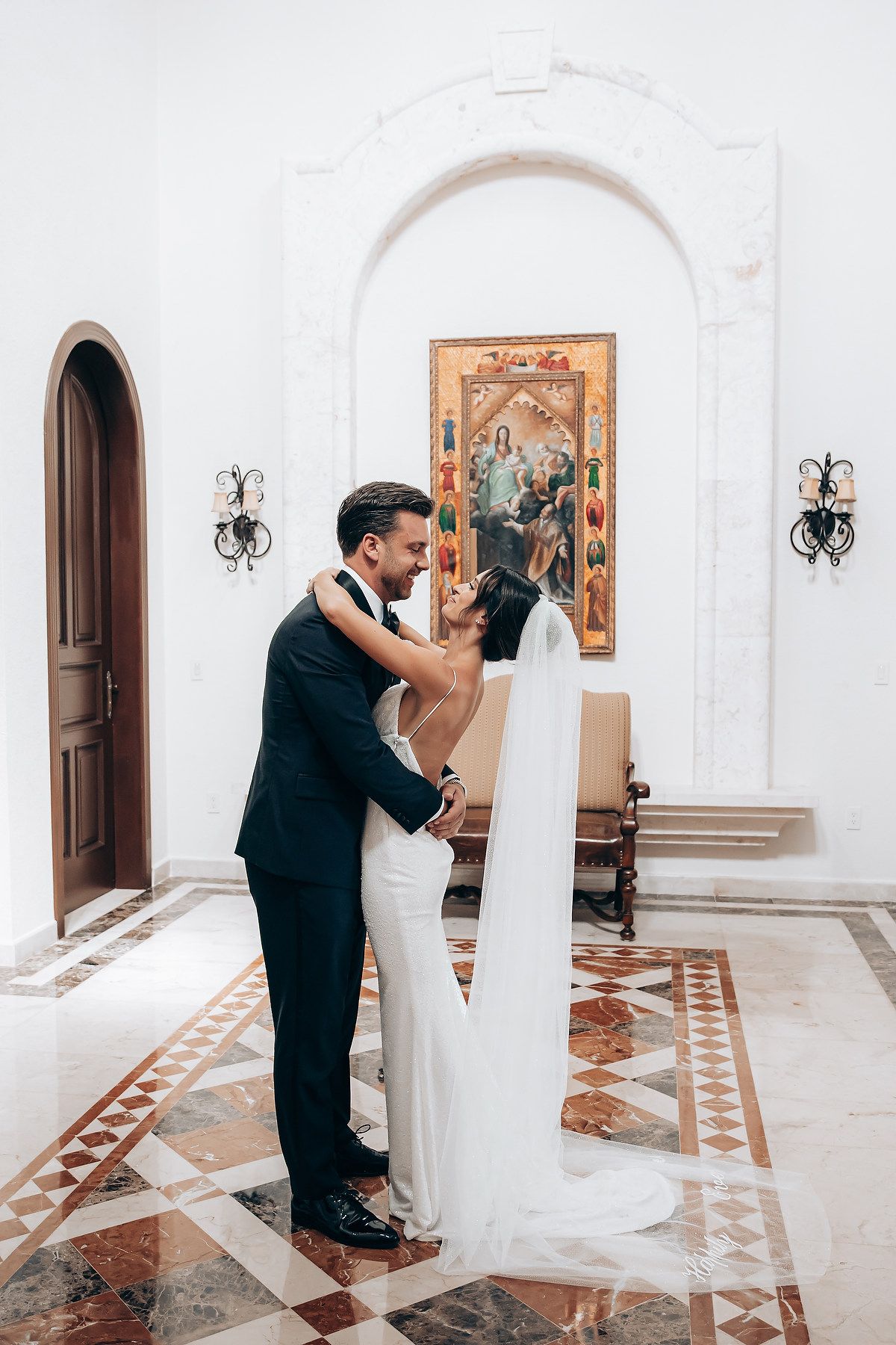 A couple dances together in an elegant white hallway with arched doorways and decorative wall art.