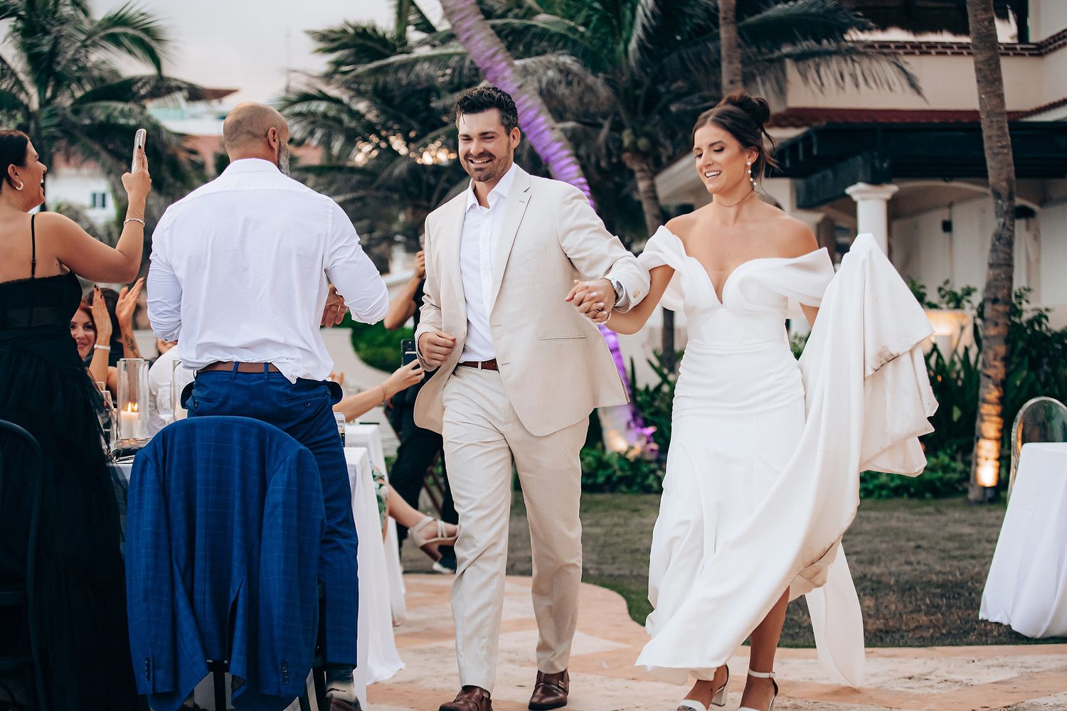 Wedding party dancing and celebrating at an outdoor tropical reception with palm trees in the background.