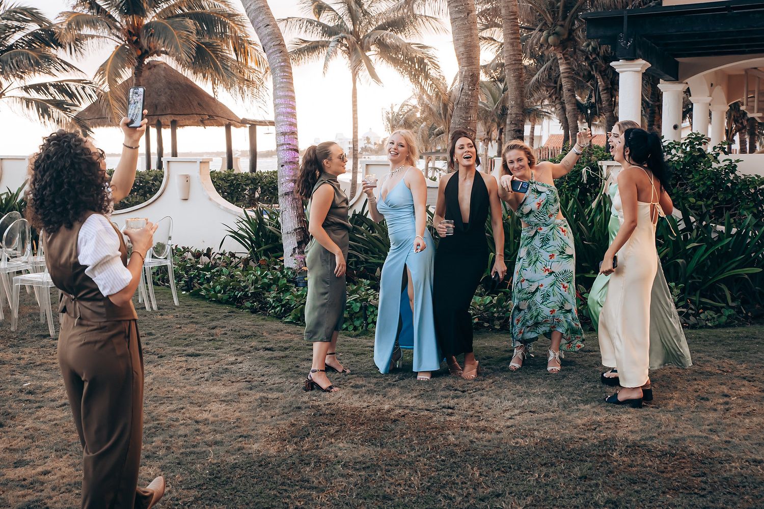 A group of people in formal attire gather under palm trees at a tropical outdoor event.