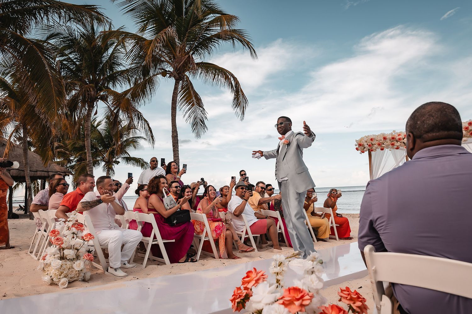 A wedding ceremony takes place outdoors under palm trees with guests seated in white chairs and coral floral decorations.