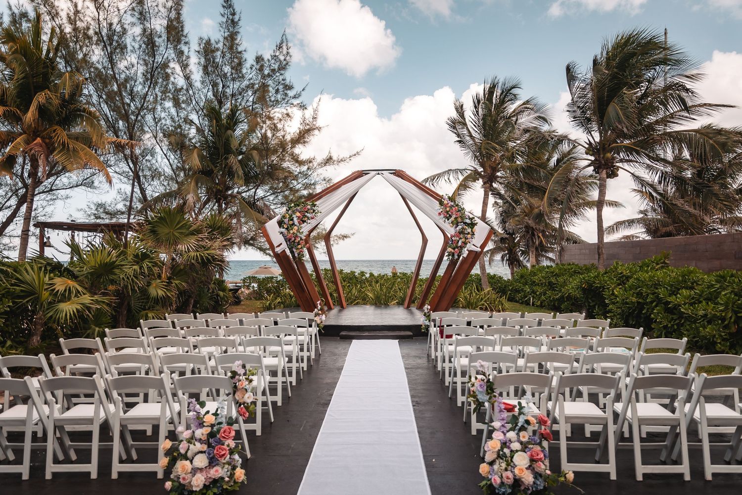Beachside wedding ceremony setup with white chairs and palm trees under a bright blue sky.