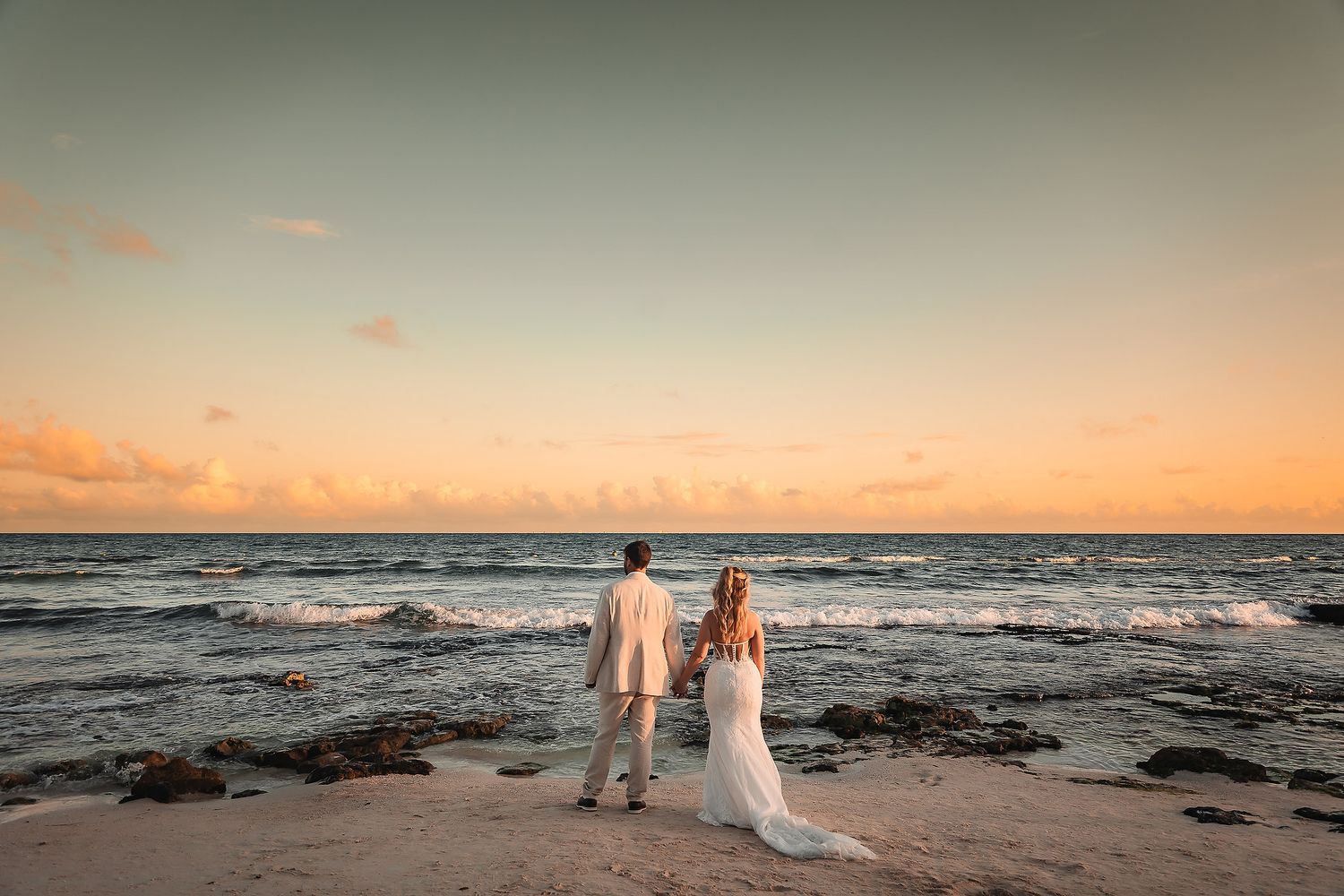 Wedding couple strolls on sandy beach during golden hour with waves crashing behind them.