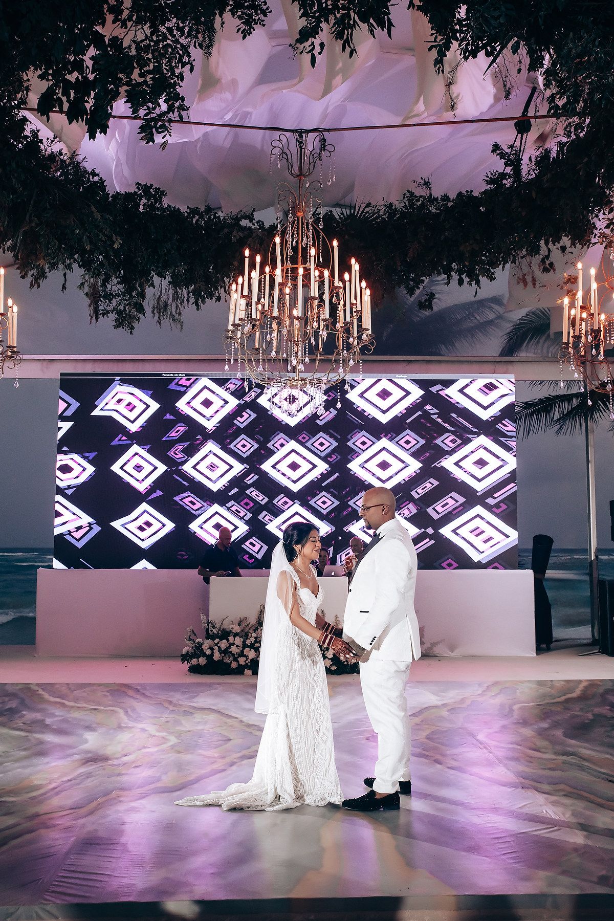 Wedding couple dancing on illuminated floor with geometric pattern backdrop and candlelit chandeliers overhead.
