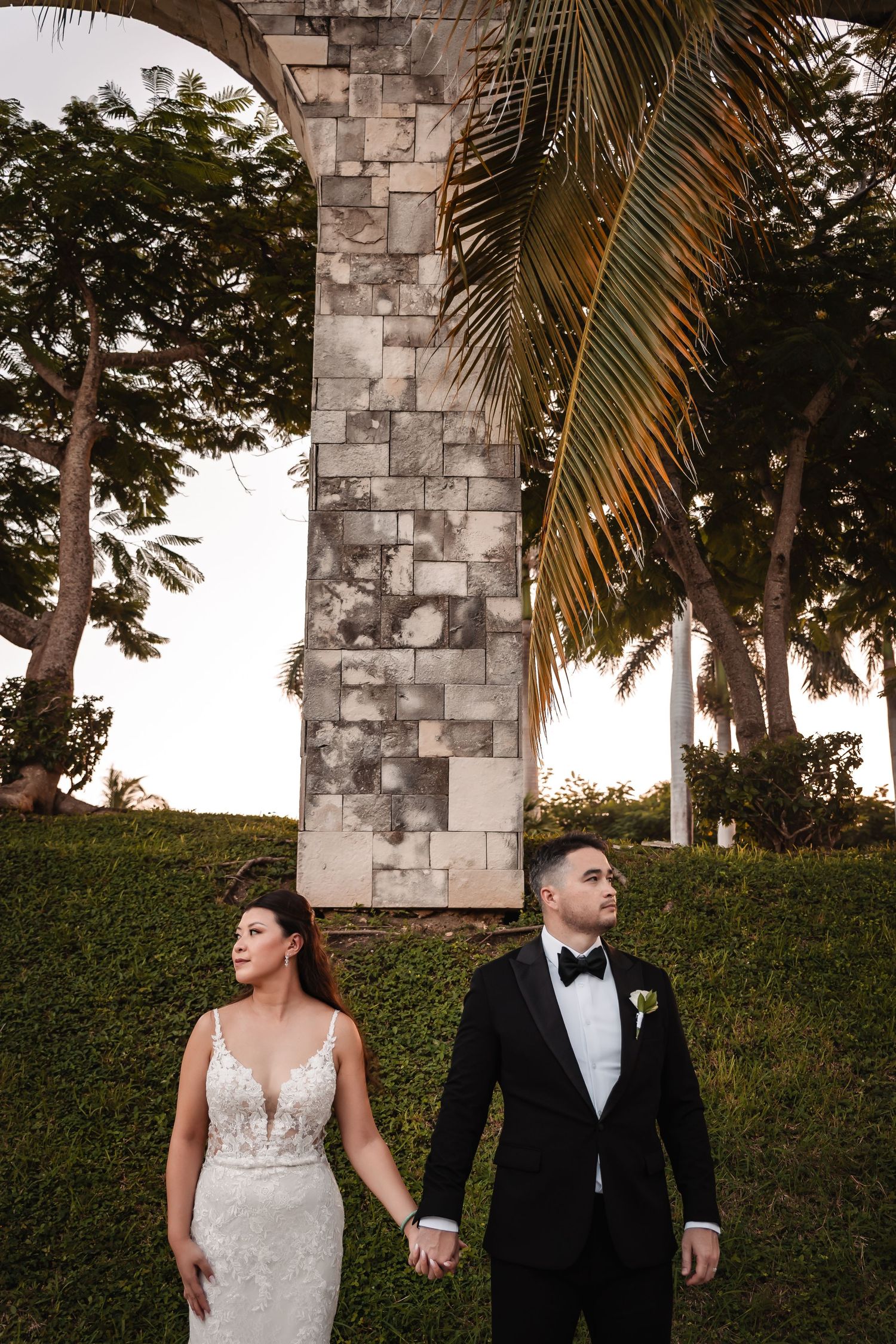 Dramatic wedding portrait with couple holding hands beneath stone architecture and palms.