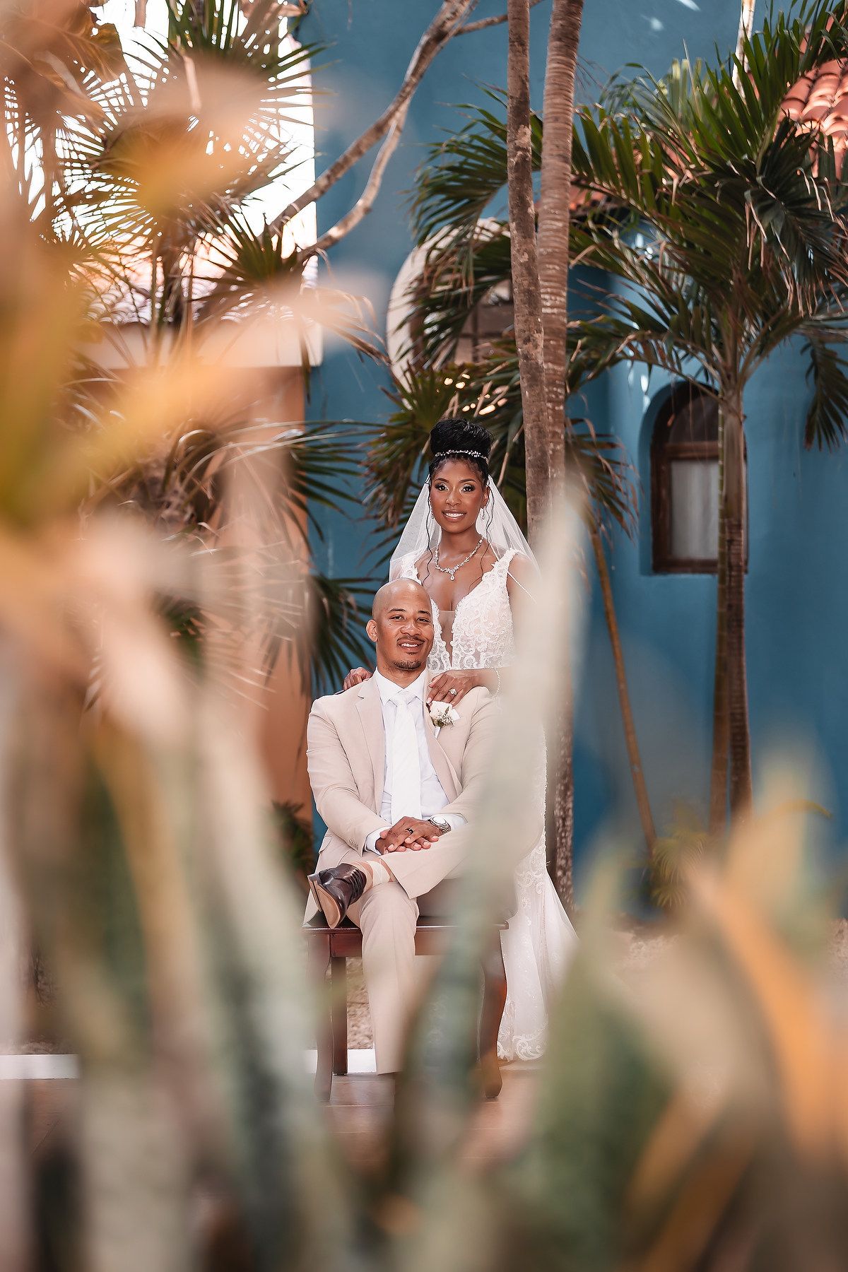 A romantic wedding portrait framed by palm trees against a bright blue wall.