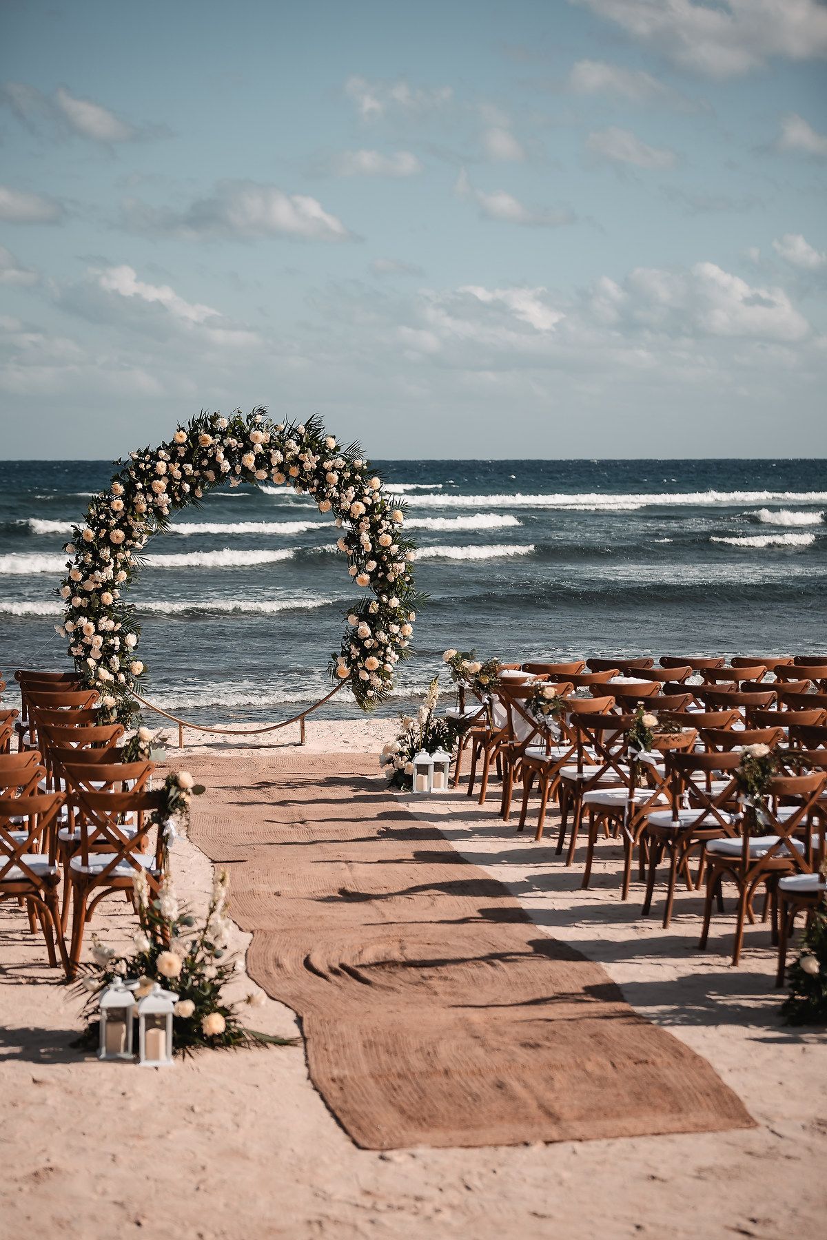 Beach wedding venue with wooden chairs and floral arch overlooking the ocean at sunset.