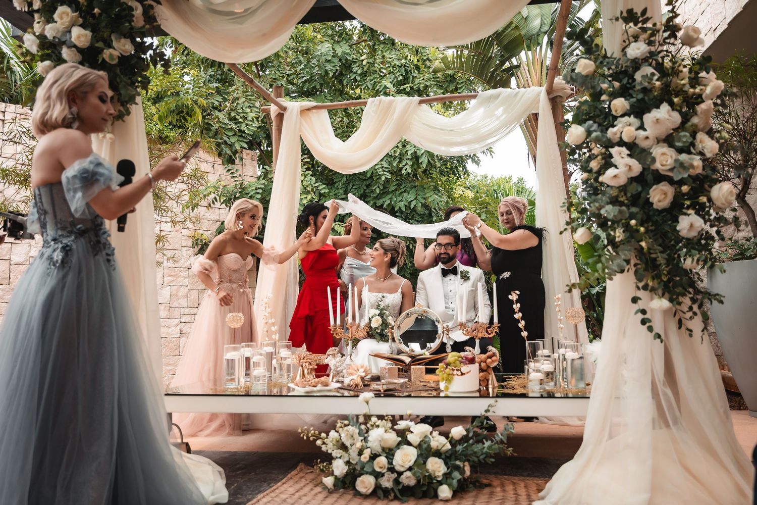 Wedding party gathers around an elegantly decorated table under a floral arch with draped fabric.