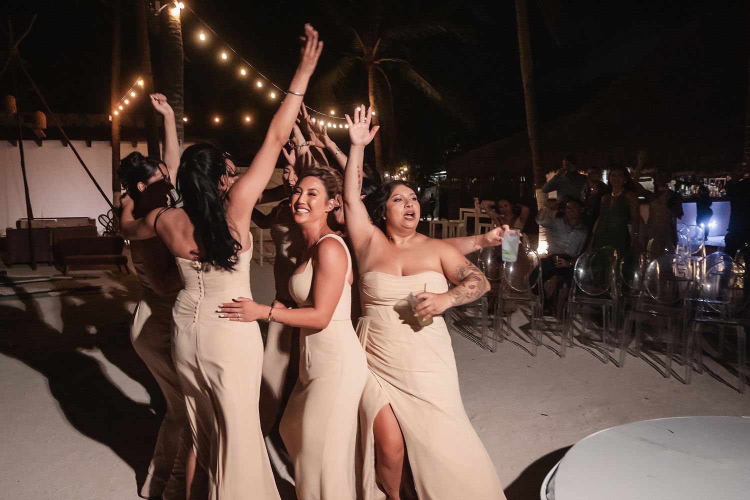 Wedding party celebrates under string lights on a beach at night.