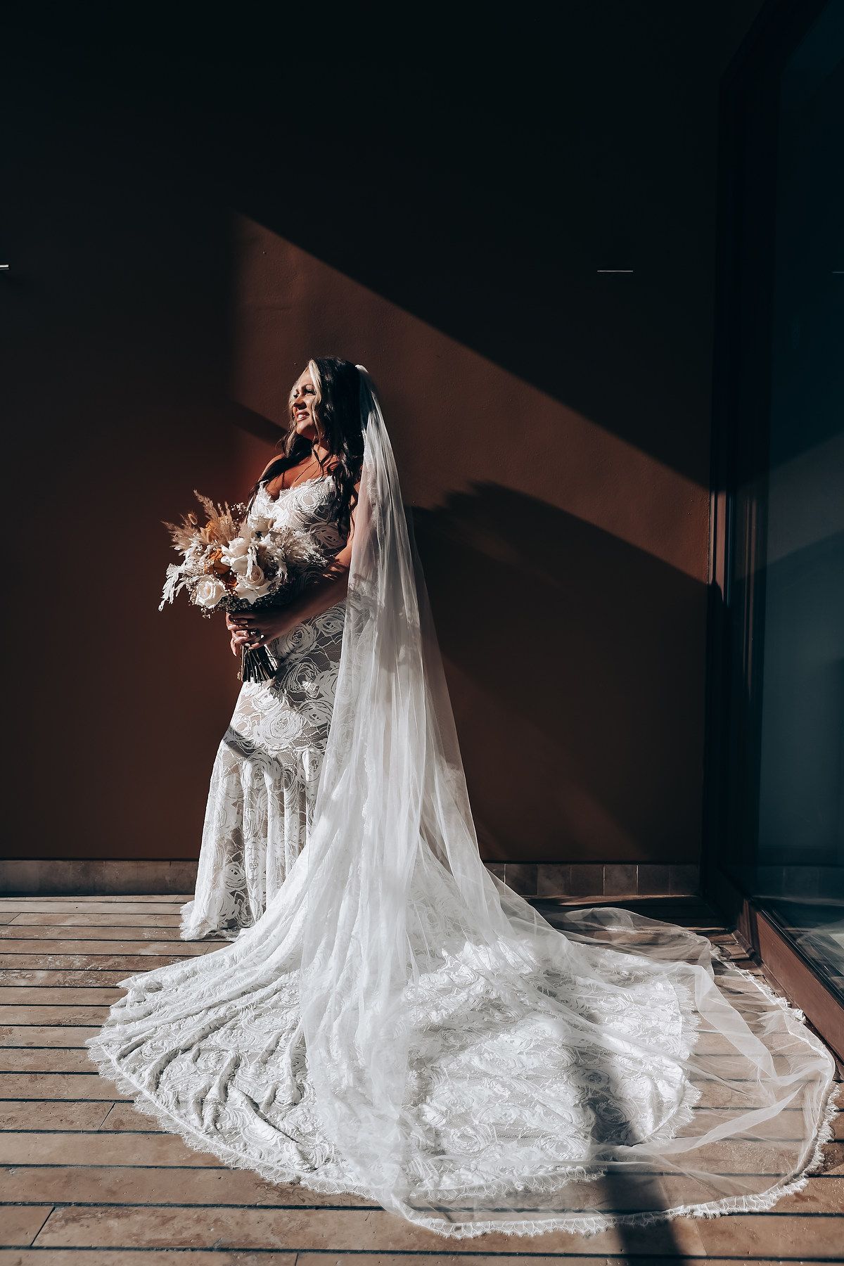 A bride in a flowing lace wedding gown with a dramatic cathedral veil poses on a wooden floor in dramatic lighting.