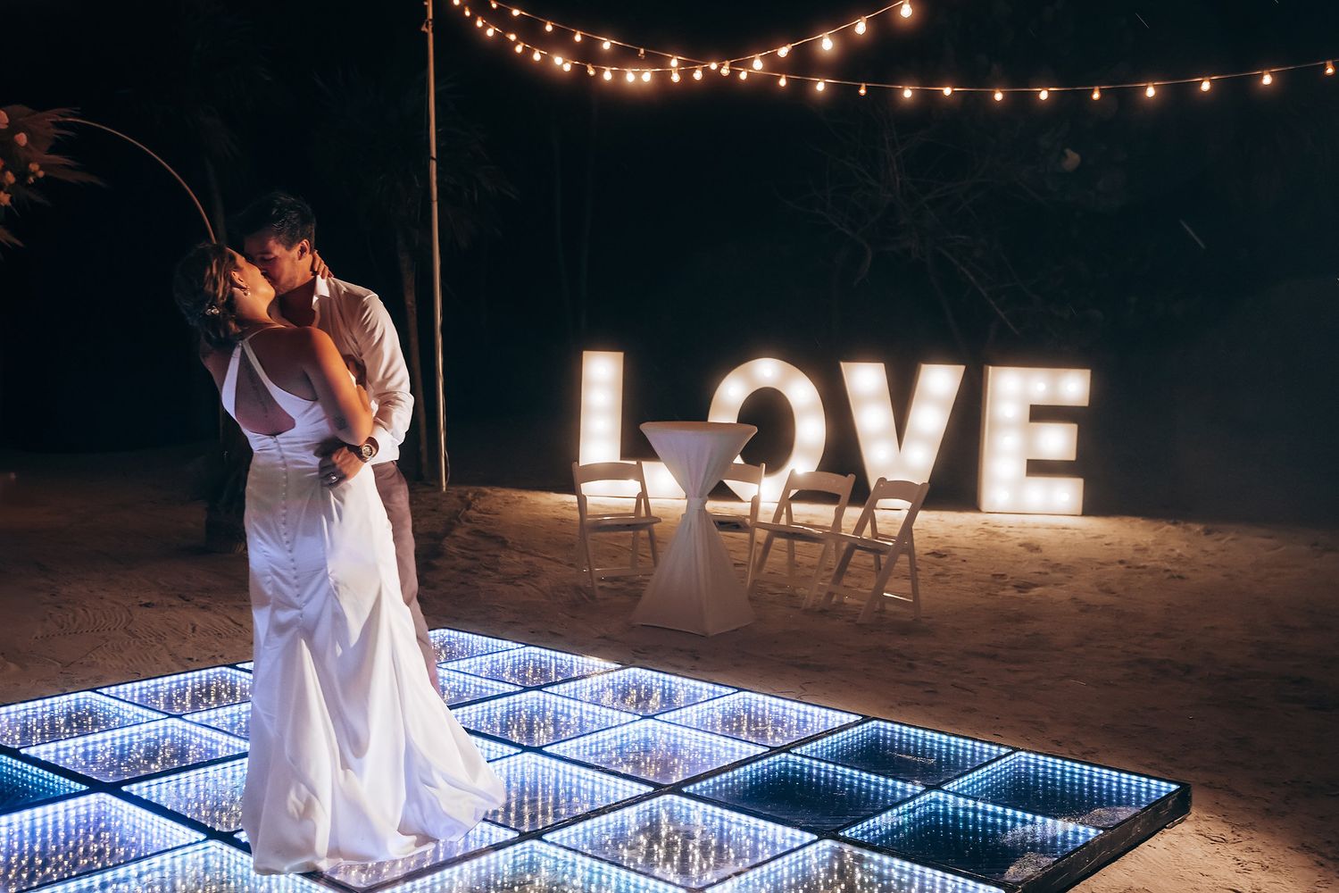 Wedding couple dancing on an illuminated LED dance floor under string lights at night.