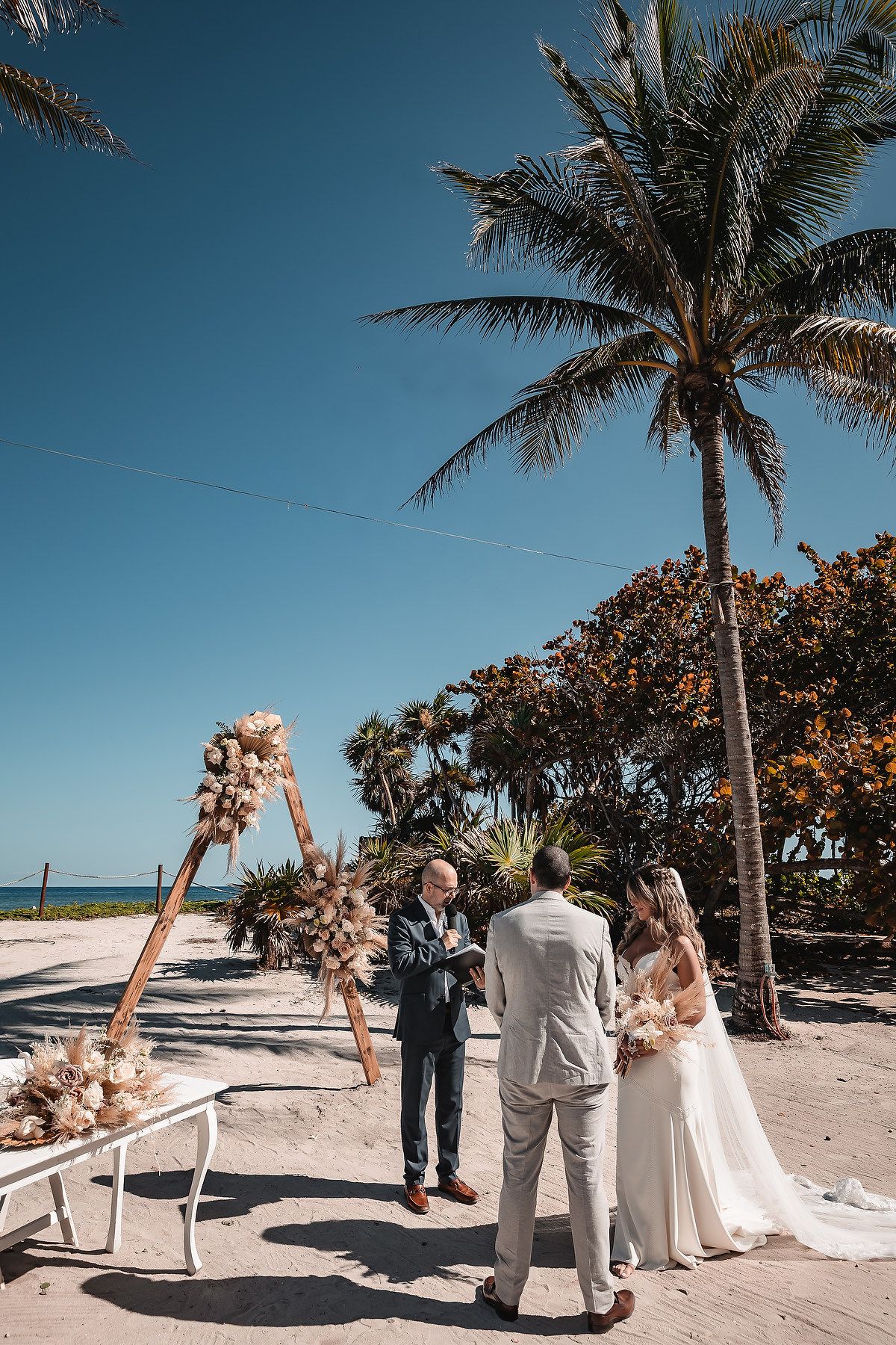 Beachfront wedding ceremony setup with palm trees and floral-adorned wooden arch against blue sky.