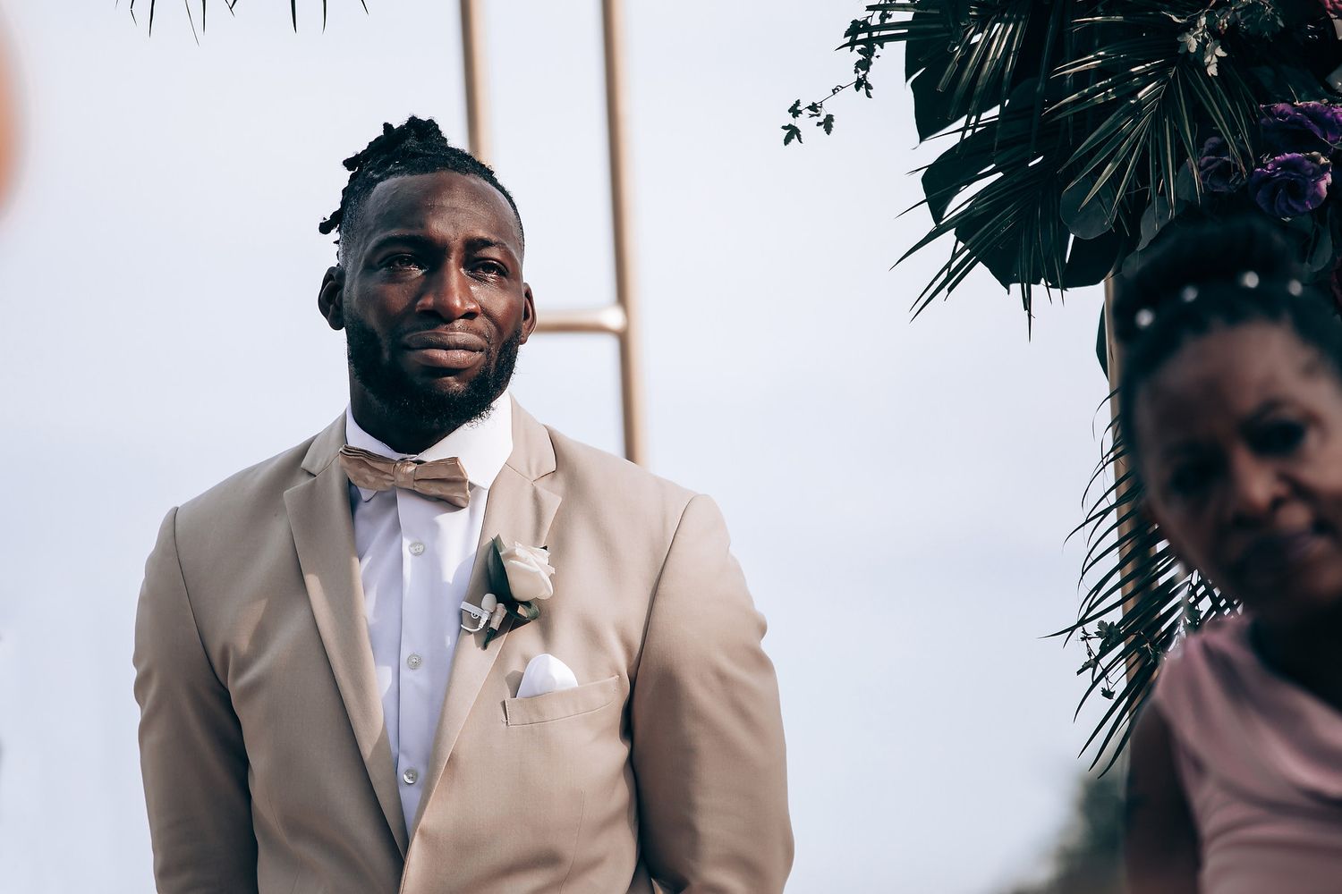 Wedding party members in beige suits stand together during a tropical beach ceremony.