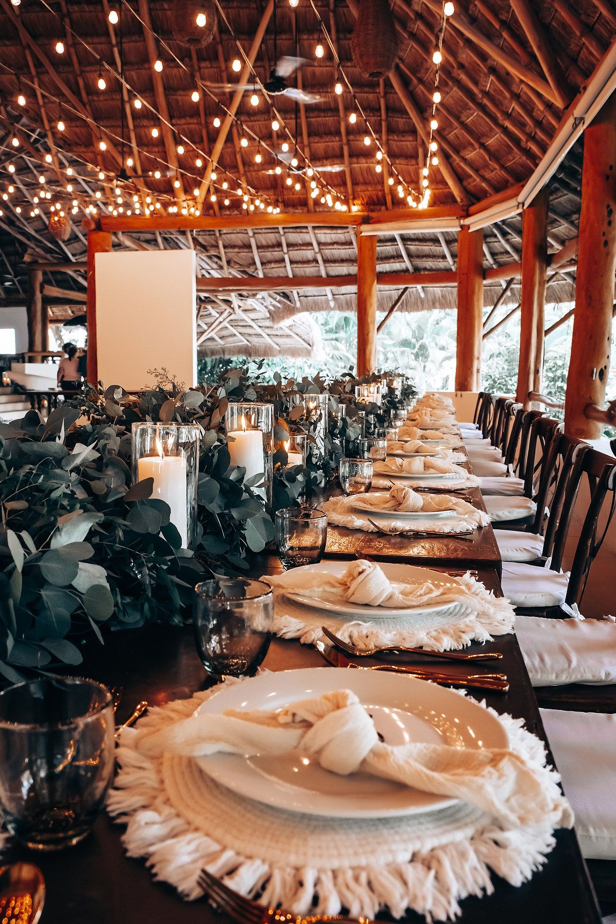 Long banquet tables set with elegant white linens and place settings under wooden beams and warm lighting.