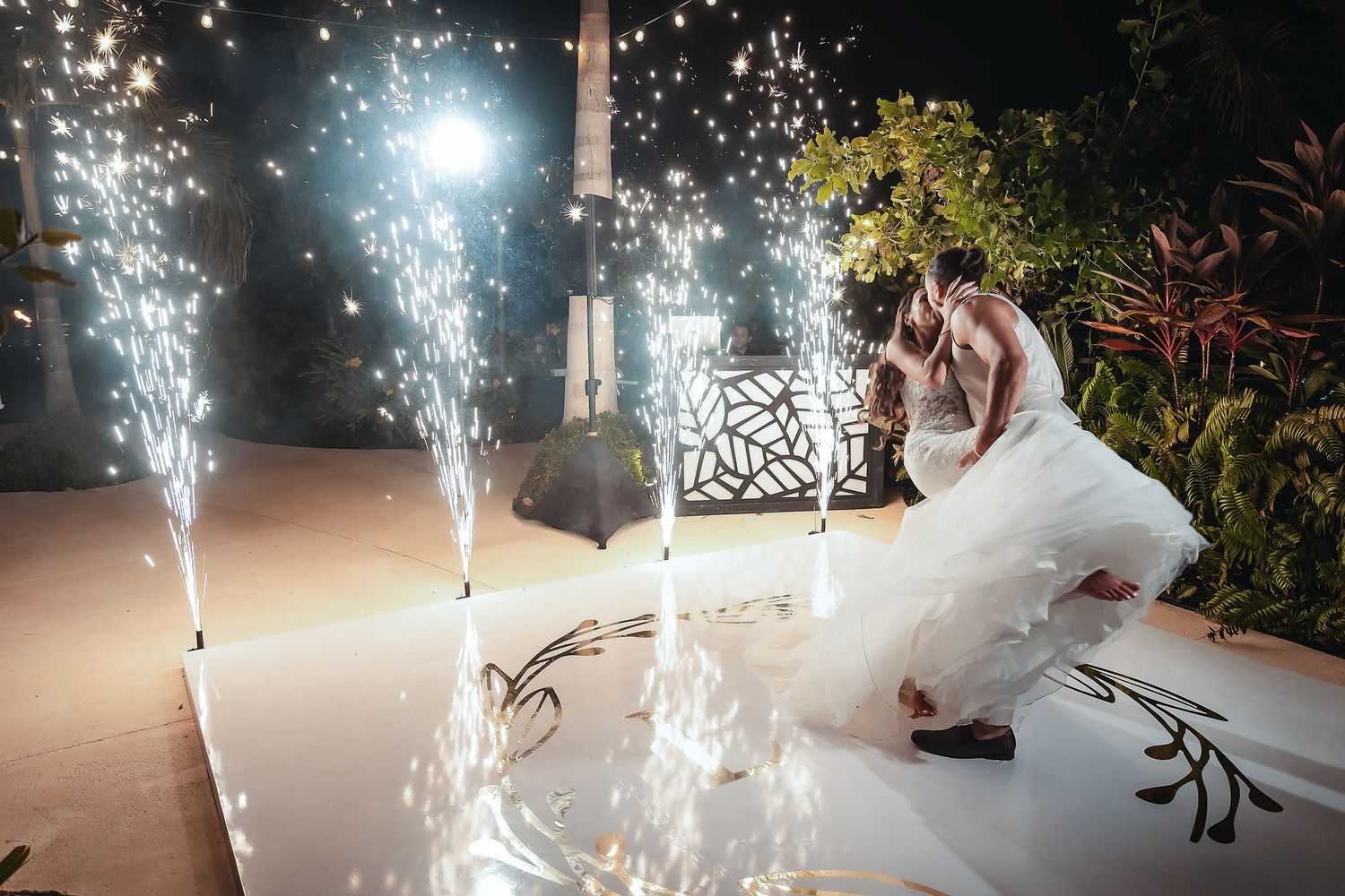 Bride and groom share a romantic kiss on a white dance floor surrounded by sparkling wedding fireworks at night.