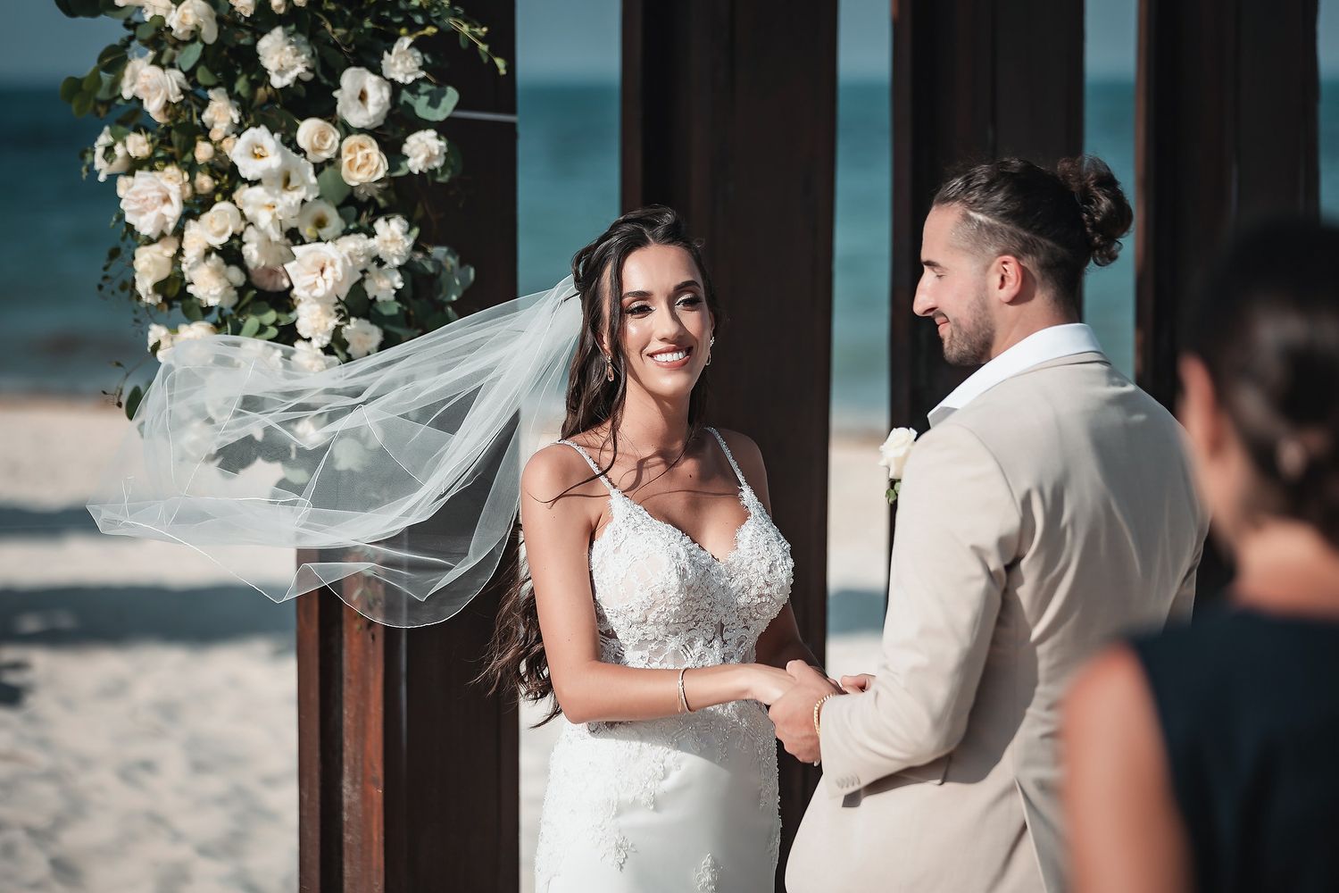 Bride and groom exchange vows at a beach wedding ceremony with white floral arch, bride wearing veil and lace gown.