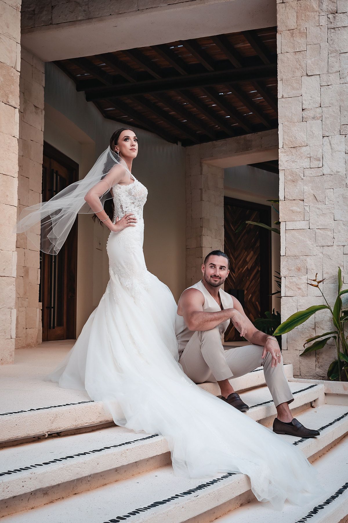 Bride in white mermaid gown with veil stands on steps while groom in tan suit sits beside her at outdoor venue.