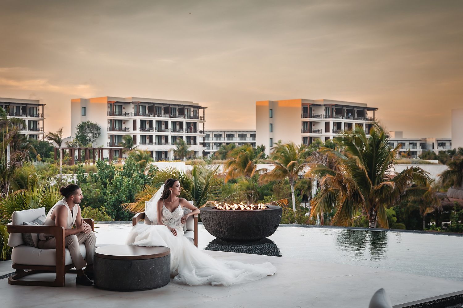 Bride and groom sit by a fire pit at a luxury resort pool during a golden sunset wedding celebration.