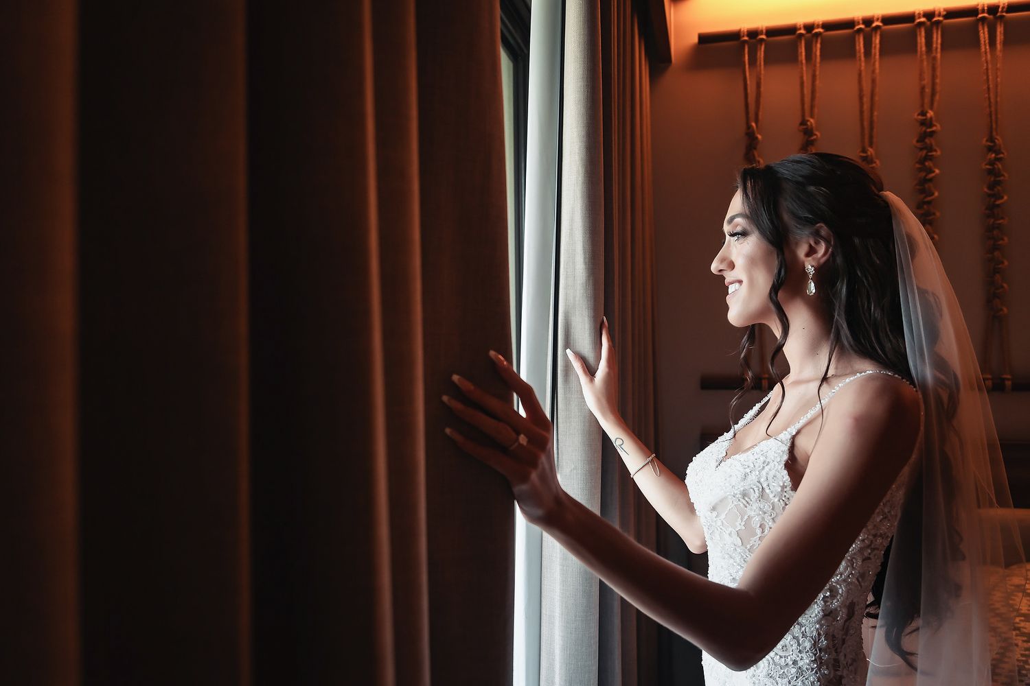 Bride in white wedding dress and veil gazing out window through curtains on her wedding day.