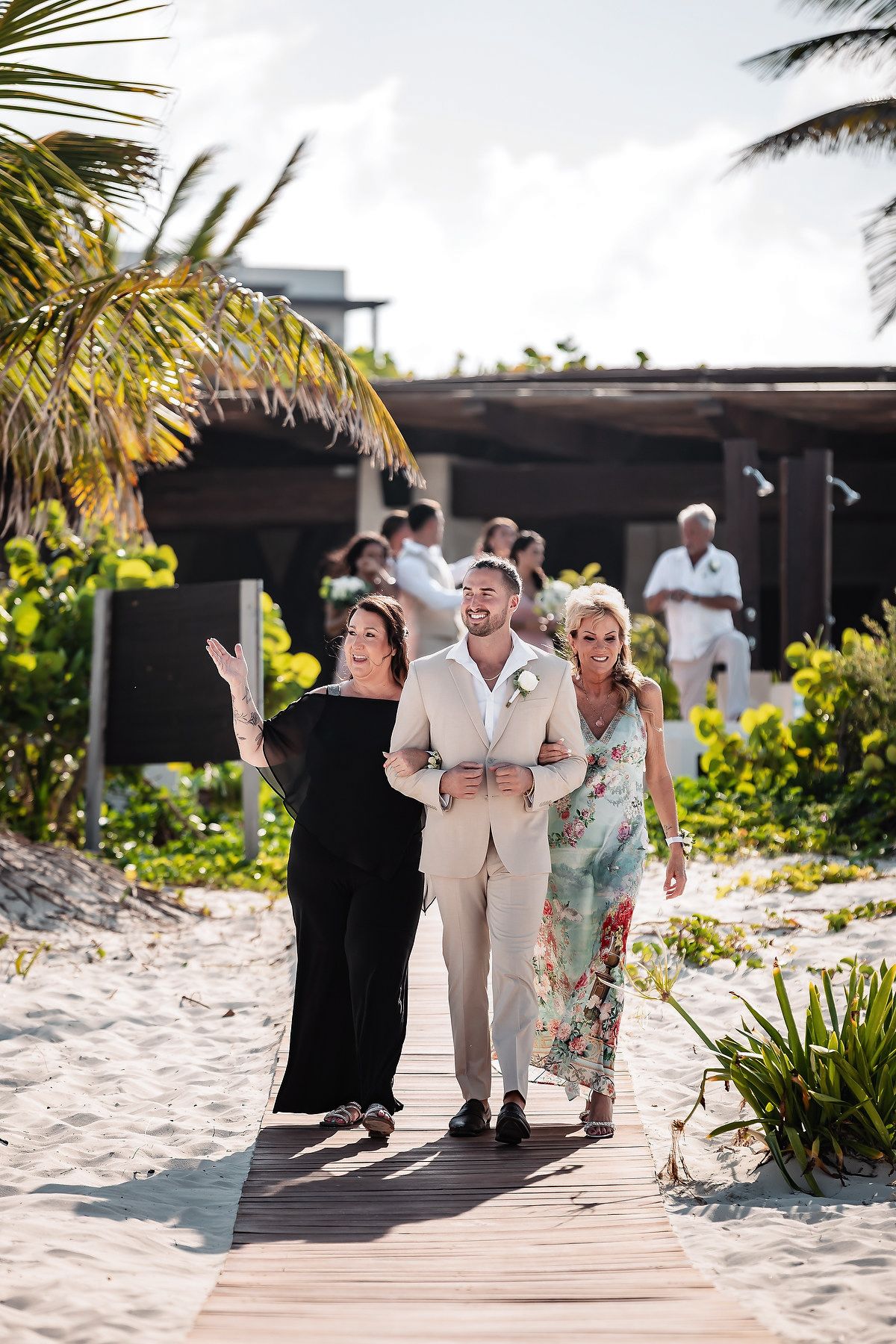 Groom in beige suit walks with two women on a wooden boardwalk at a tropical beach wedding venue.