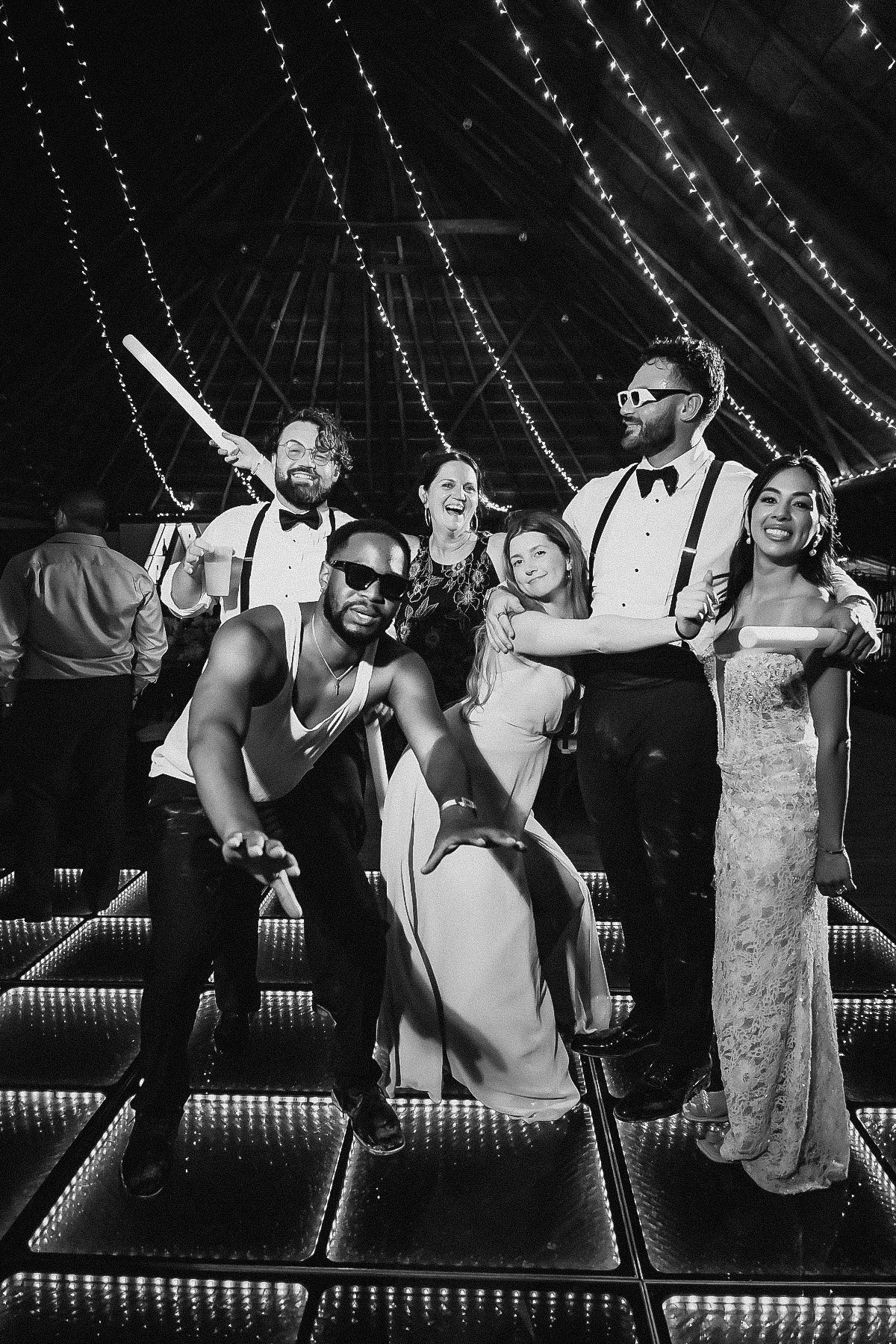 Black and white photo of a joyful wedding party posing on a dance floor under twinkling lights in a barn venue.